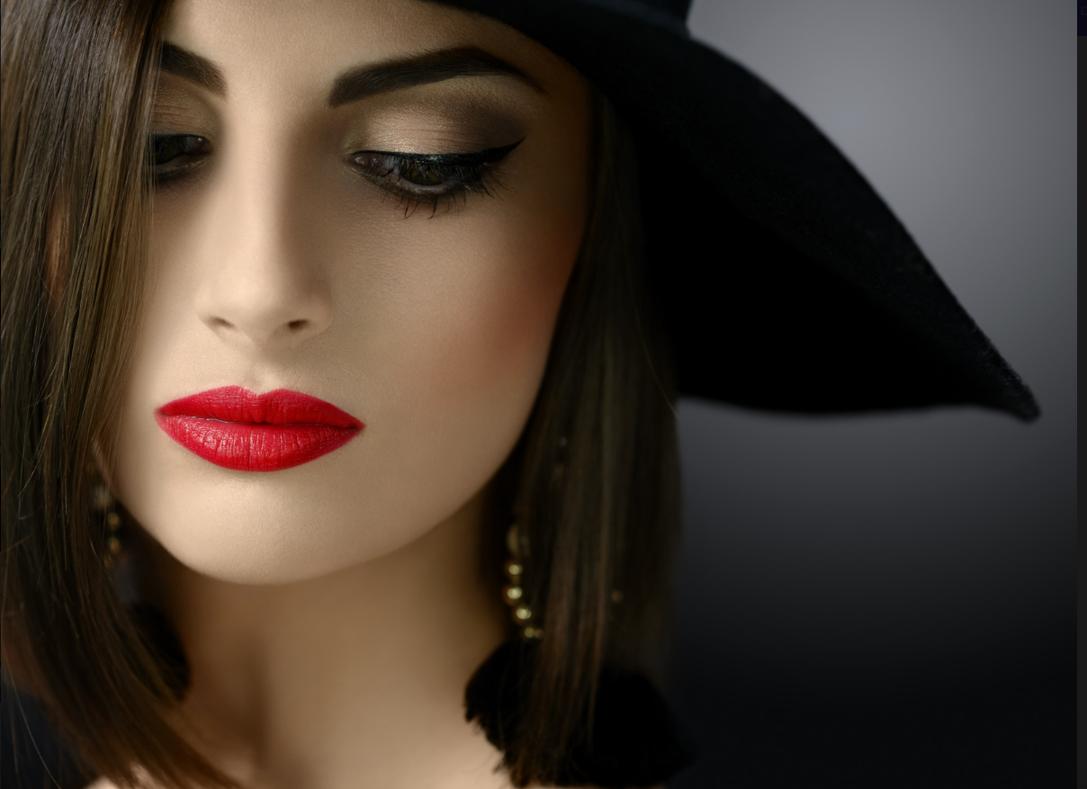 Ritratto di una donna con trucco elegante, cappello nero e orecchini pendenti, con trucco occhi e labbra distinti, sfondo scuro.