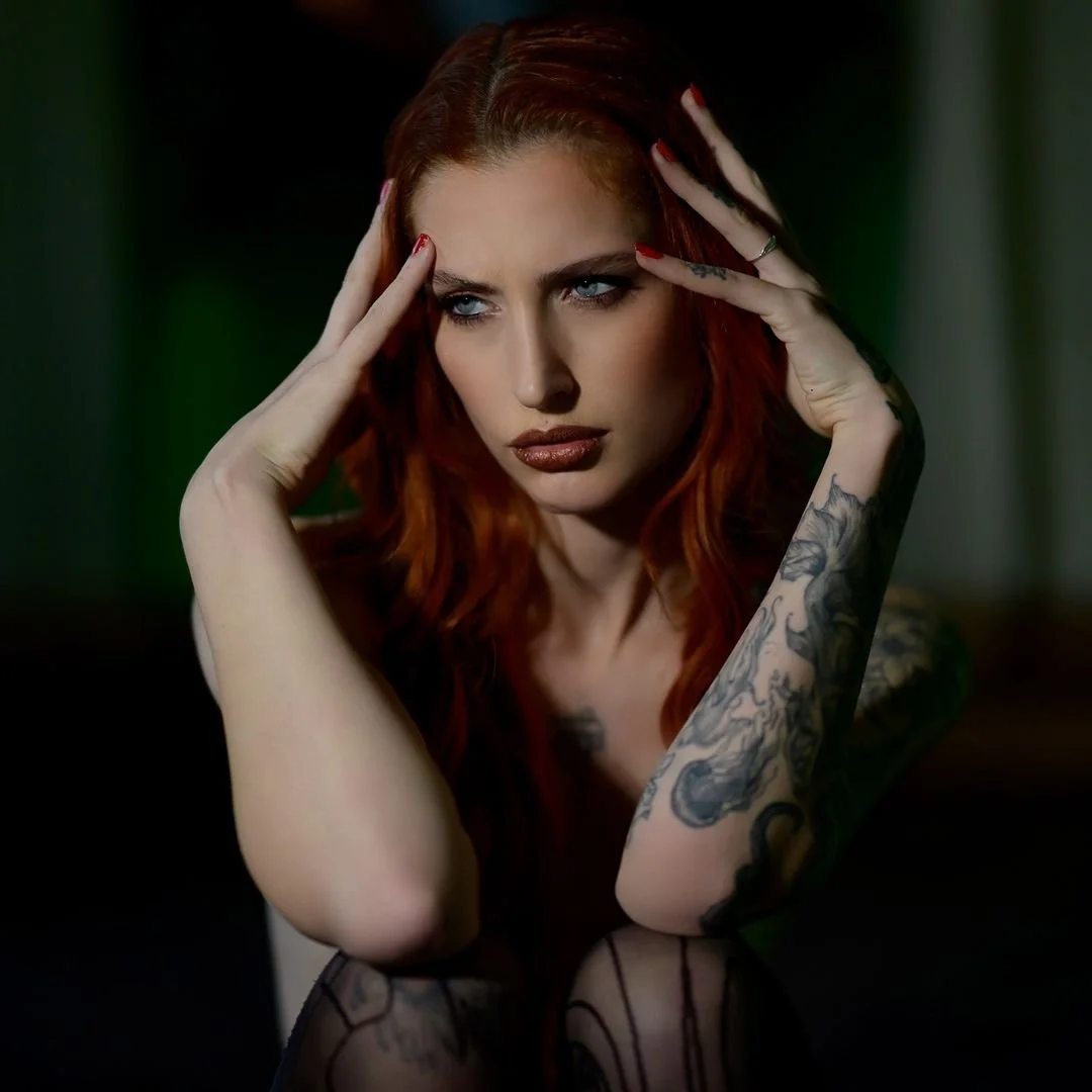 Una donna con i capelli rossi, trucco scuro, posa con le mani sulla testa, mostra tatuaggi sulle braccia e un'espressione intensa in uno sfondo scuro.