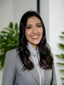 Donna sorridente con capelli scuri, indossa un abito grigio e sfondo con piante verdi.