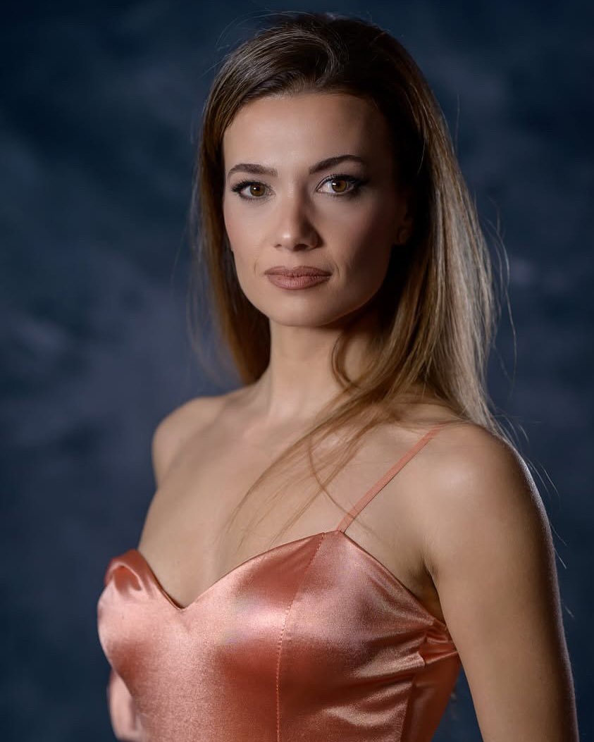 Una donna con capelli castani, occhi chiari e trucco naturale, indossa un vestito in raso di colore rosa pesca, con spalline sottili, posa contro uno sfondo scuro.