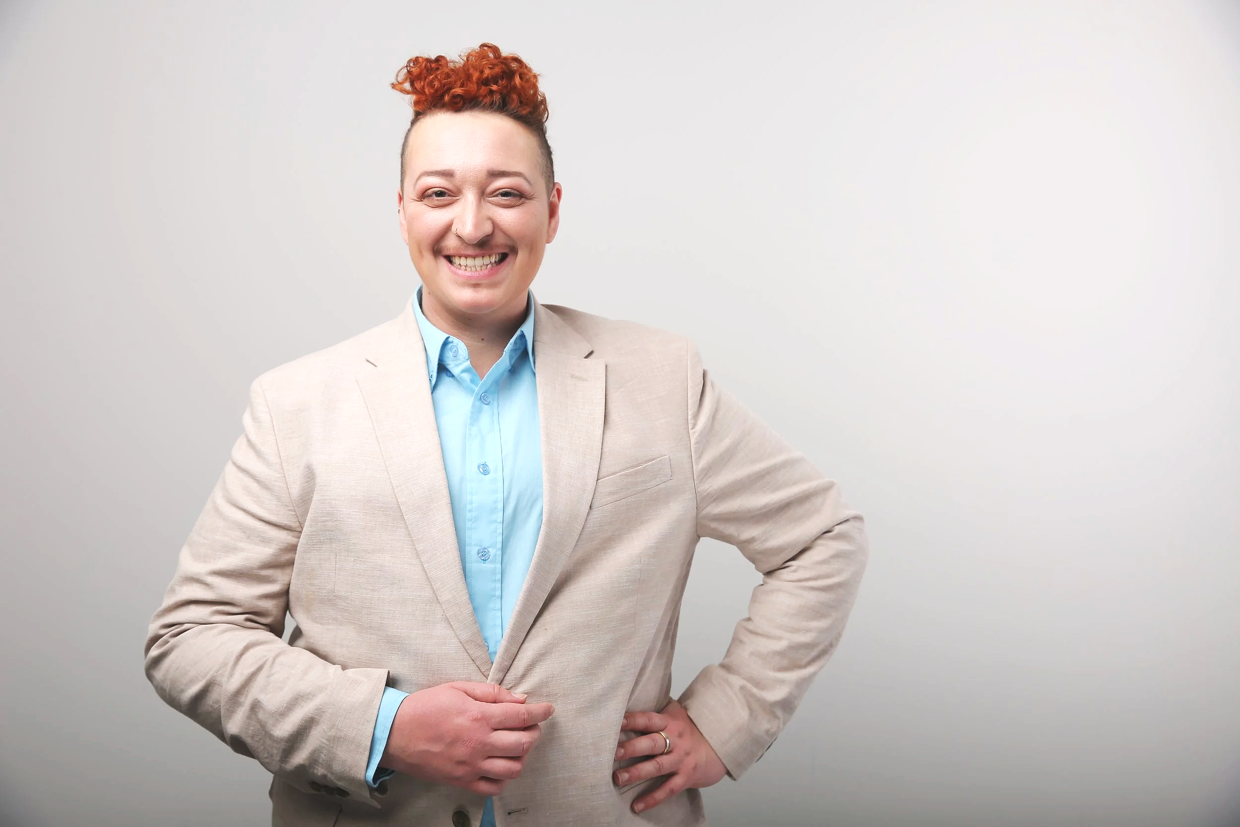 Eine Person mit roten Locken, die eine beigefarbene Jacke und ein blaues Hemd trägt, lächelt vor einem weißen Hintergrund. Der linke Arm ist in die Hüfte gestemmt.