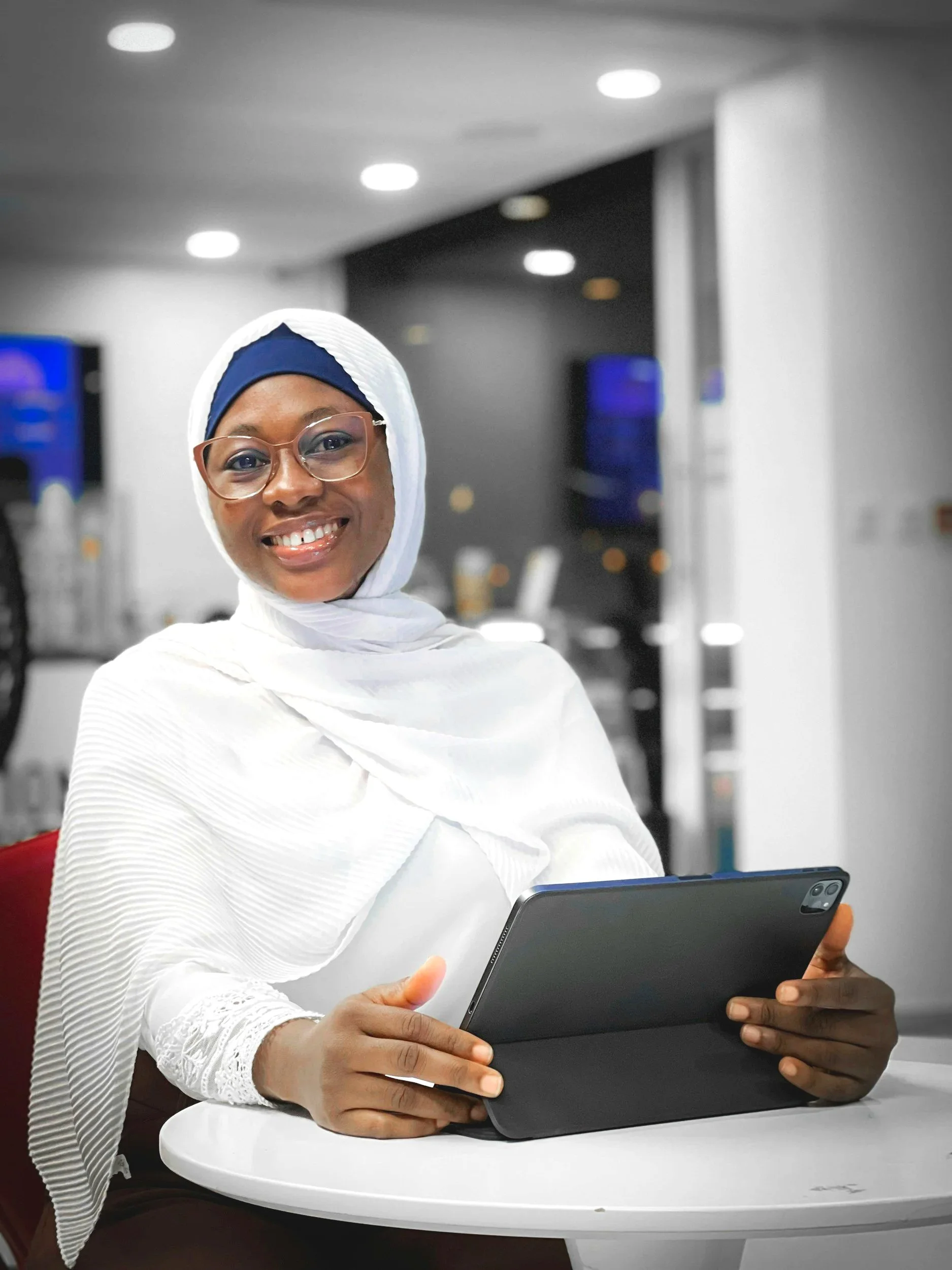 Eine Person of Color mit Brille und Kopftuch, die einen Tablet-Computer hält und in einem Büro sitzt und in die Kamera lächelt.