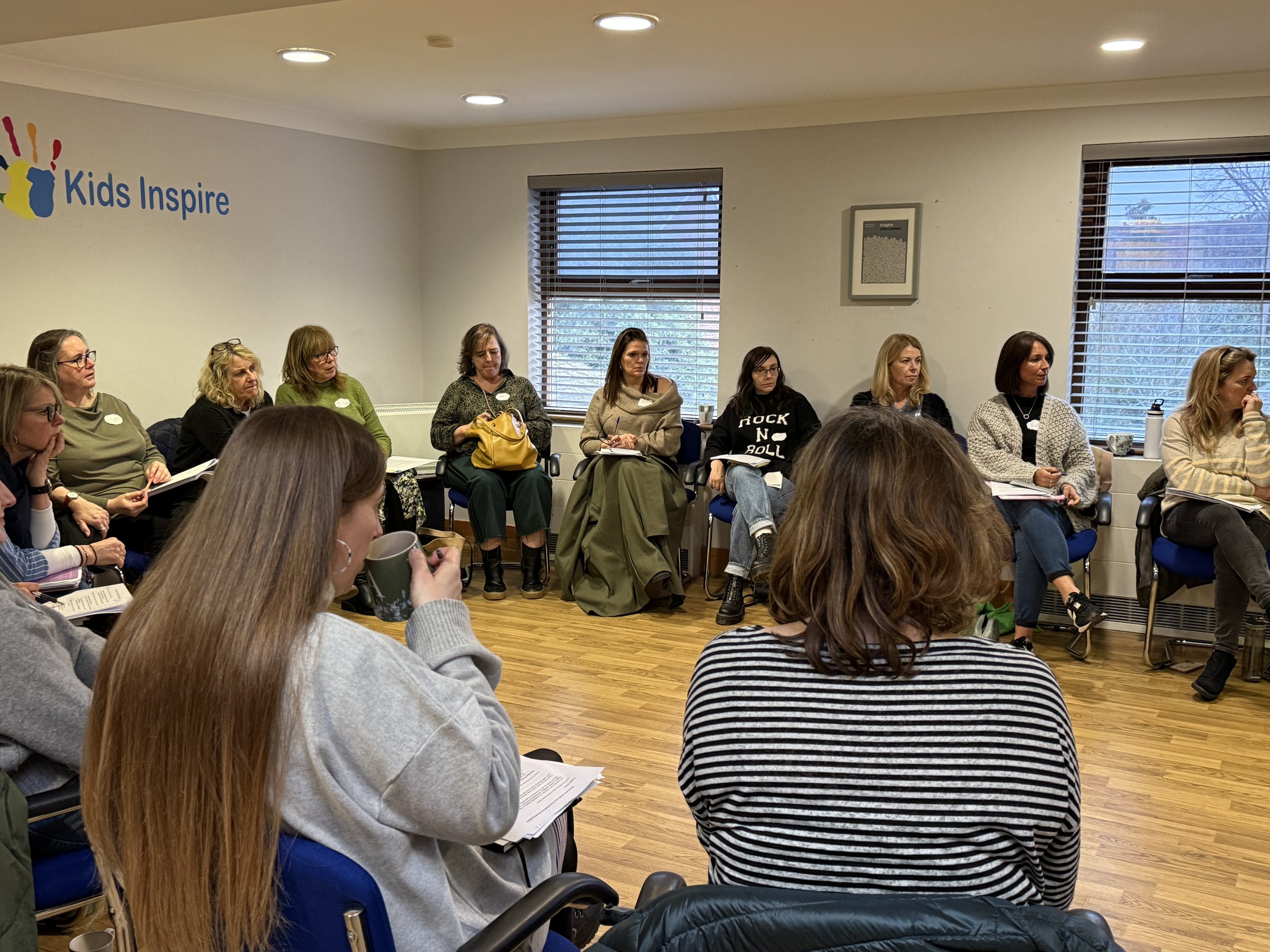 A group of women sitting in a room attending a meeting or seminar, some taking notes, with windows in the background and a wall with the words 'Kids Inspire' and a colorful handprint logo.