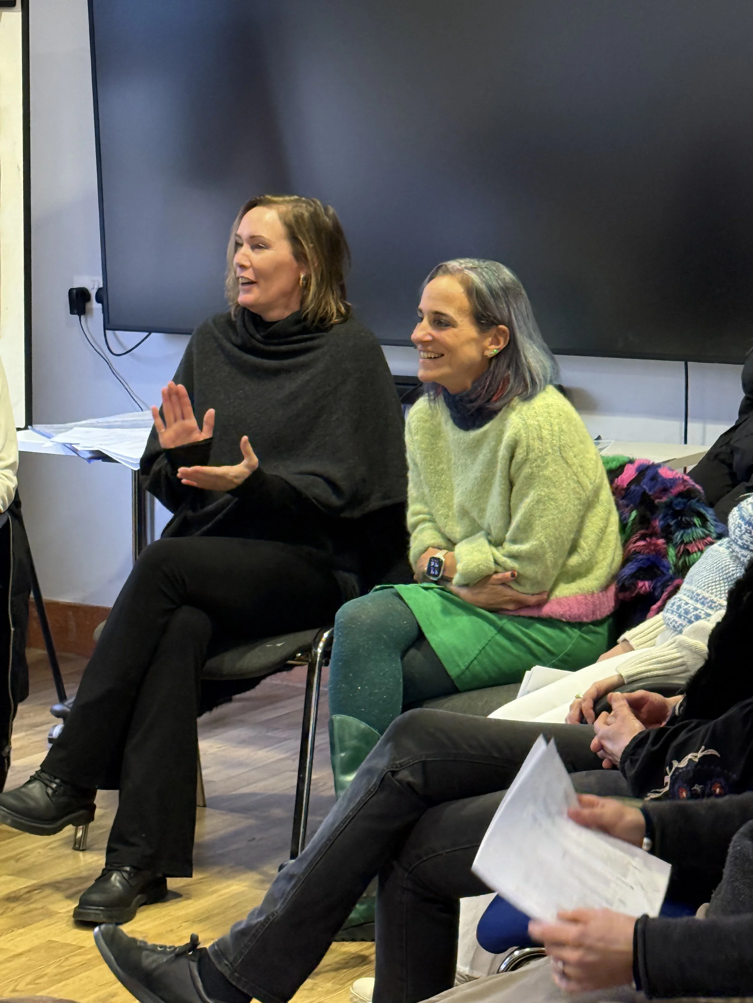 Two women sitting in a meeting, one speaking and gesturing while the other listens, with papers in the hands of nearby participants. A television is in the background.