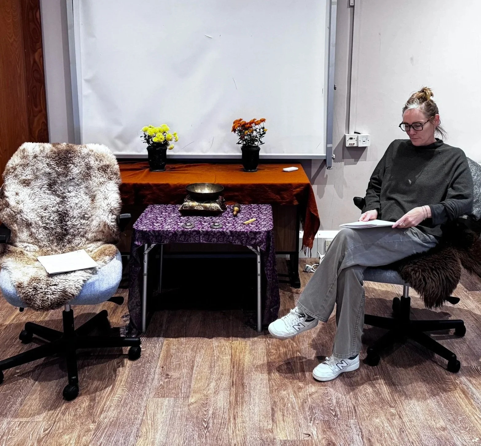 A woman sitting in a chair with a fur-covered blanket, holding papers, and a fluffy chair with a white note on it is visible. The room has a table with yellow and orange flowers, a singing bowl, and a purple cloth, with a whiteboard in the background.