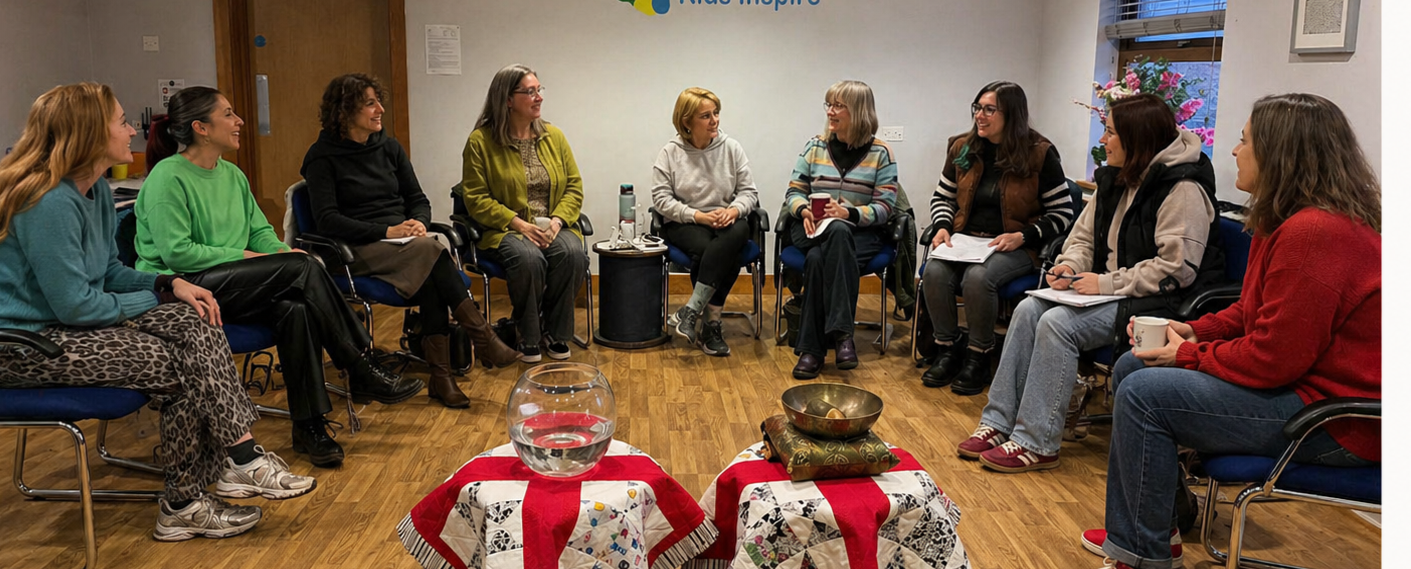 A group of women sitting in a circle in a room, engaging in a discussion. There are two tables with cloths, a fishbowl, a singing bowl, and a cushion in the center. The women are holding notebooks, mugs, and a water bottle.