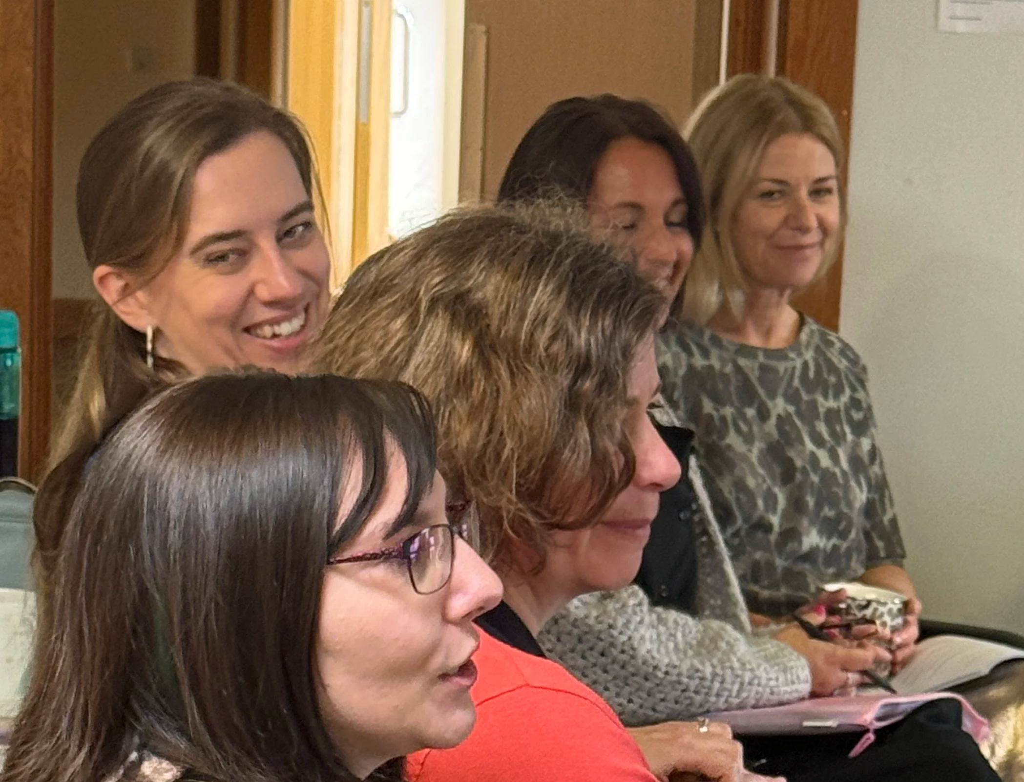 A group of women sitting in a row, some holding phones, in an indoor setting.