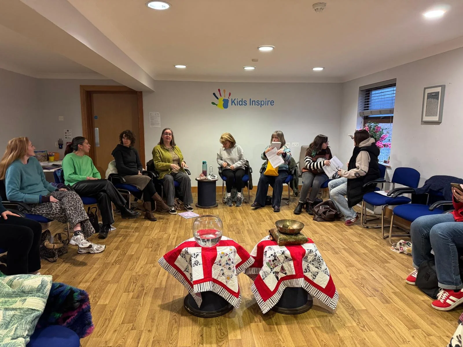Group of women sitting in a circle in a room with a sign that reads 'Kids Inspire' on the wall. Two tables and some items are in the center of the room.