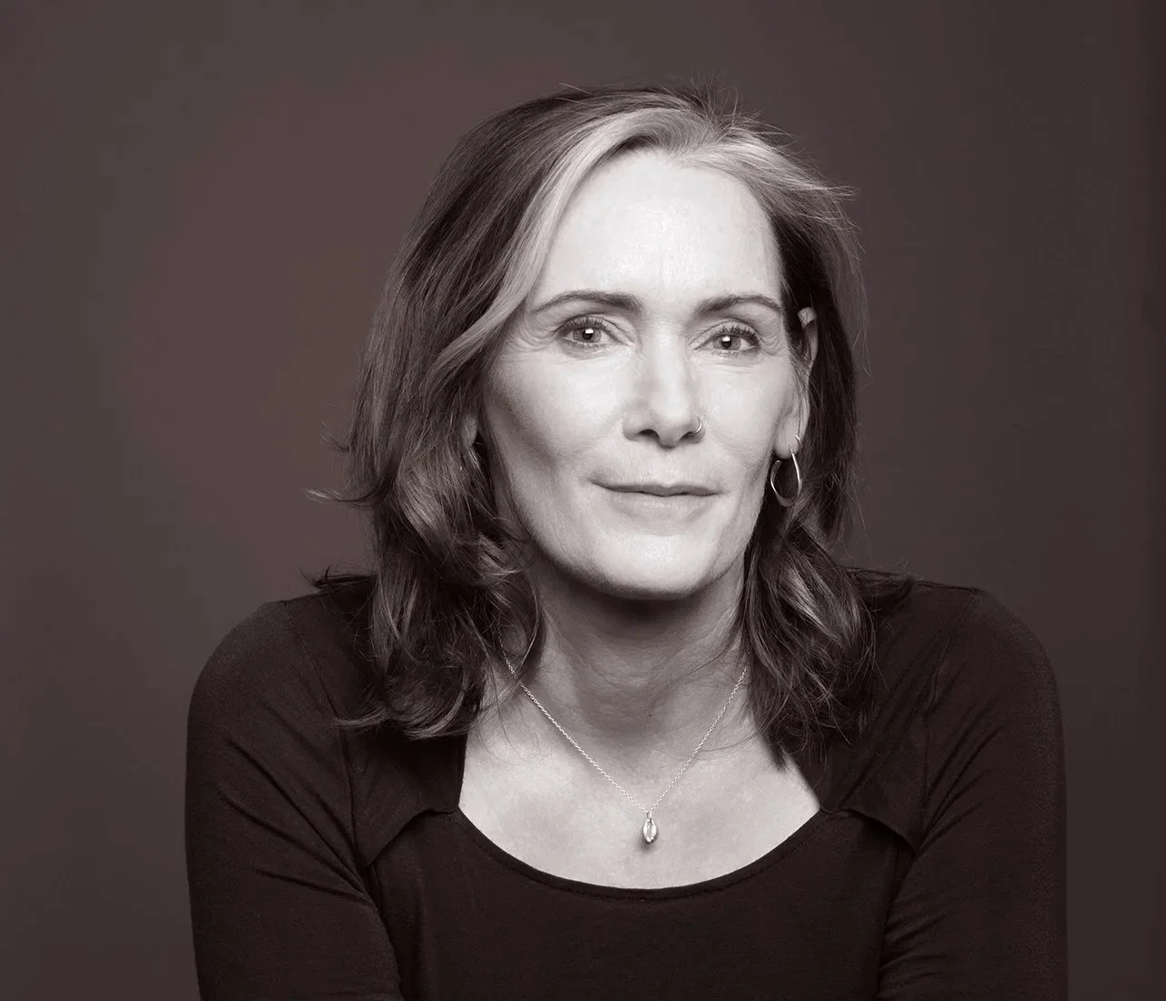 Black and white portrait of a woman with wavy shoulder-length hair, wearing a necklace and earrings, against a plain background.