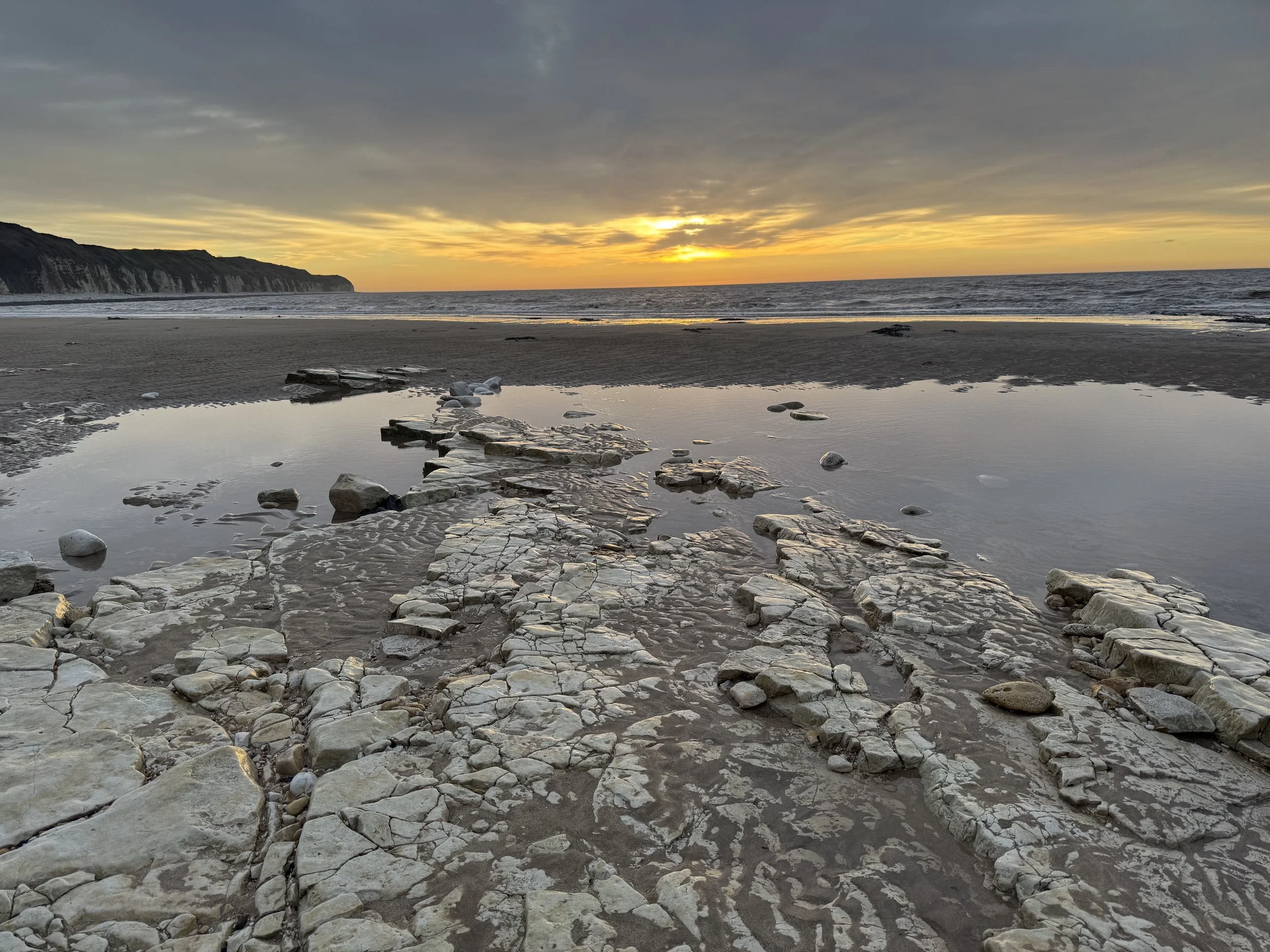 Sunset over a sandy beach with rocky formations and a calm water pool in the foreground. Cliffs in the distance under a partly cloudy sky.