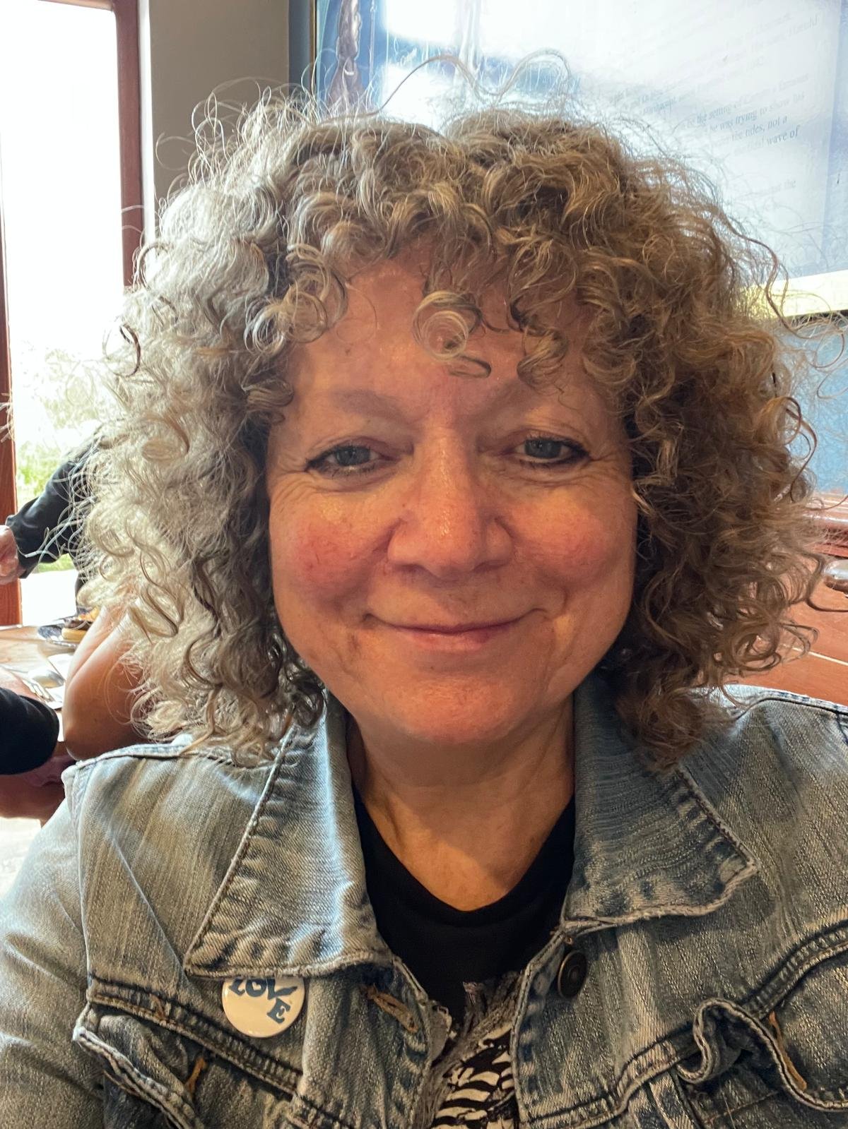 A woman with curly blonde hair smiling at the camera, wearing a denim jacket with a sticker that reads 'Vote,' sitting in a room with a window and a large screen in the background.