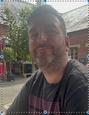 A man with short dark hair and a beard sitting outdoors near a brick building, under a tree.