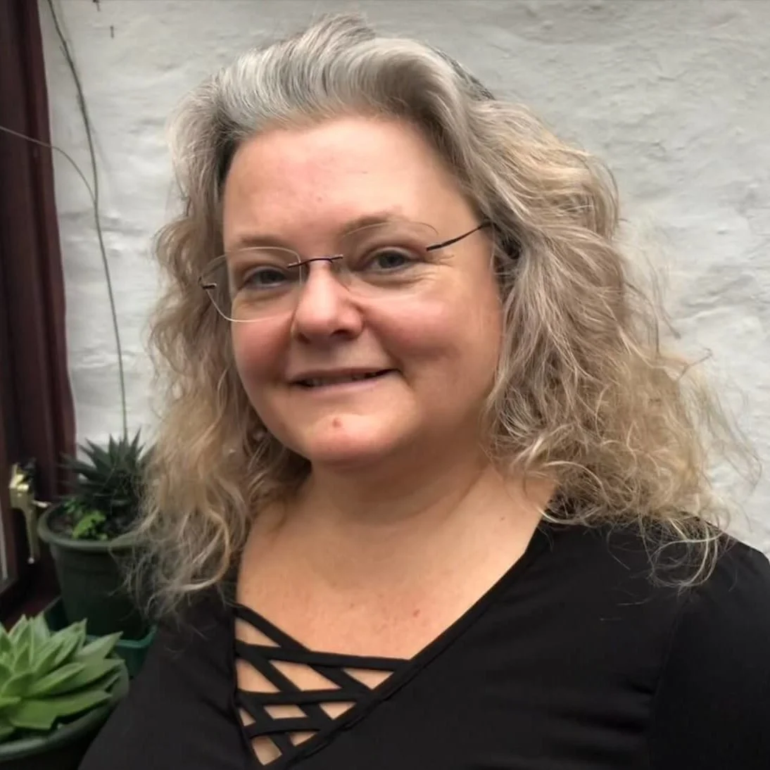 A mature woman with curly, shoulder-length gray hair, wearing glasses and a black top with a crisscross neckline, smiling at the camera. There are potted plants on a shelf behind her against a white brick wall.