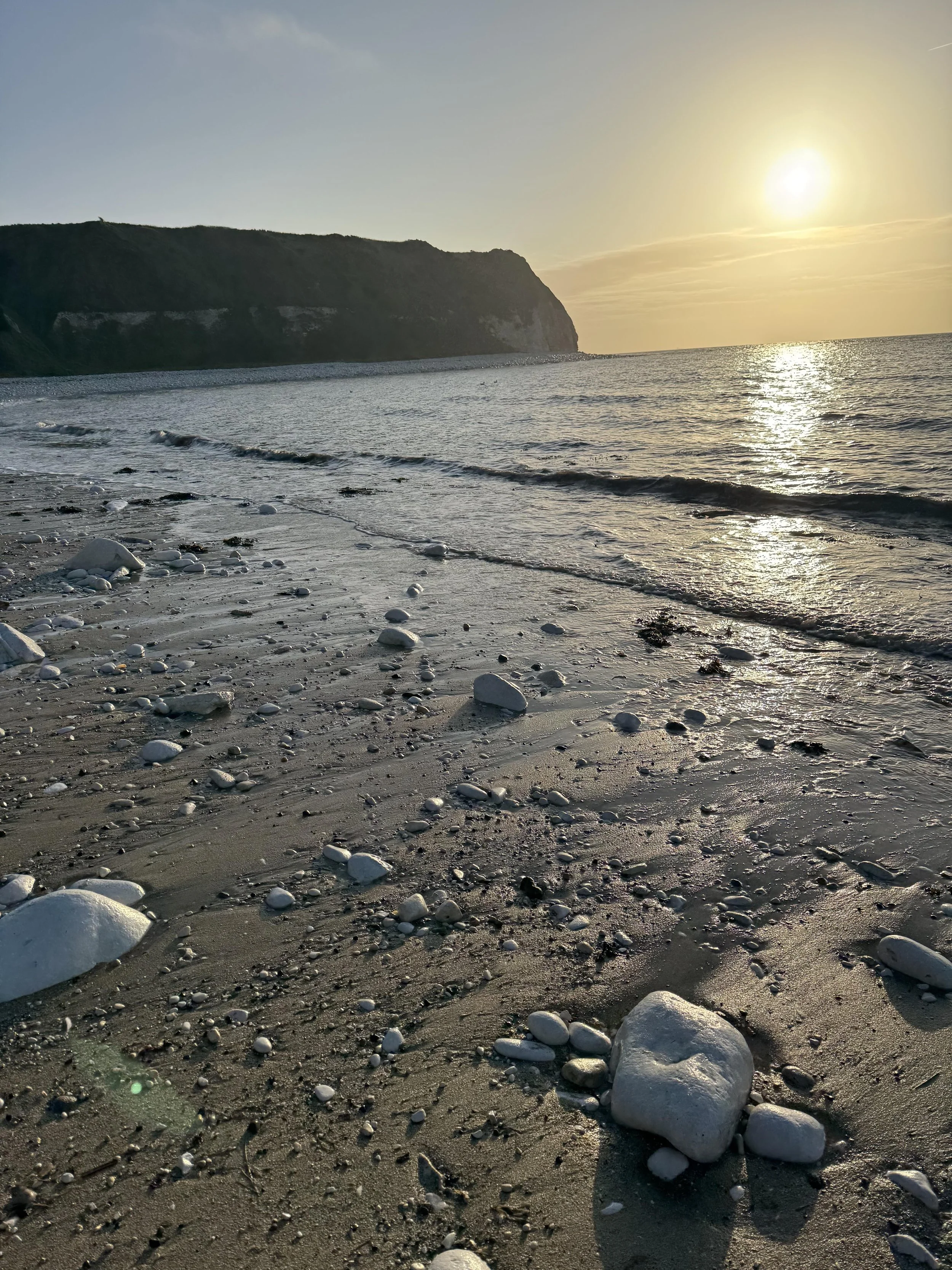A rocky beach with large white stones scattered on the sand, gentle waves, a cliff in the distance, and the sun setting on the horizon.