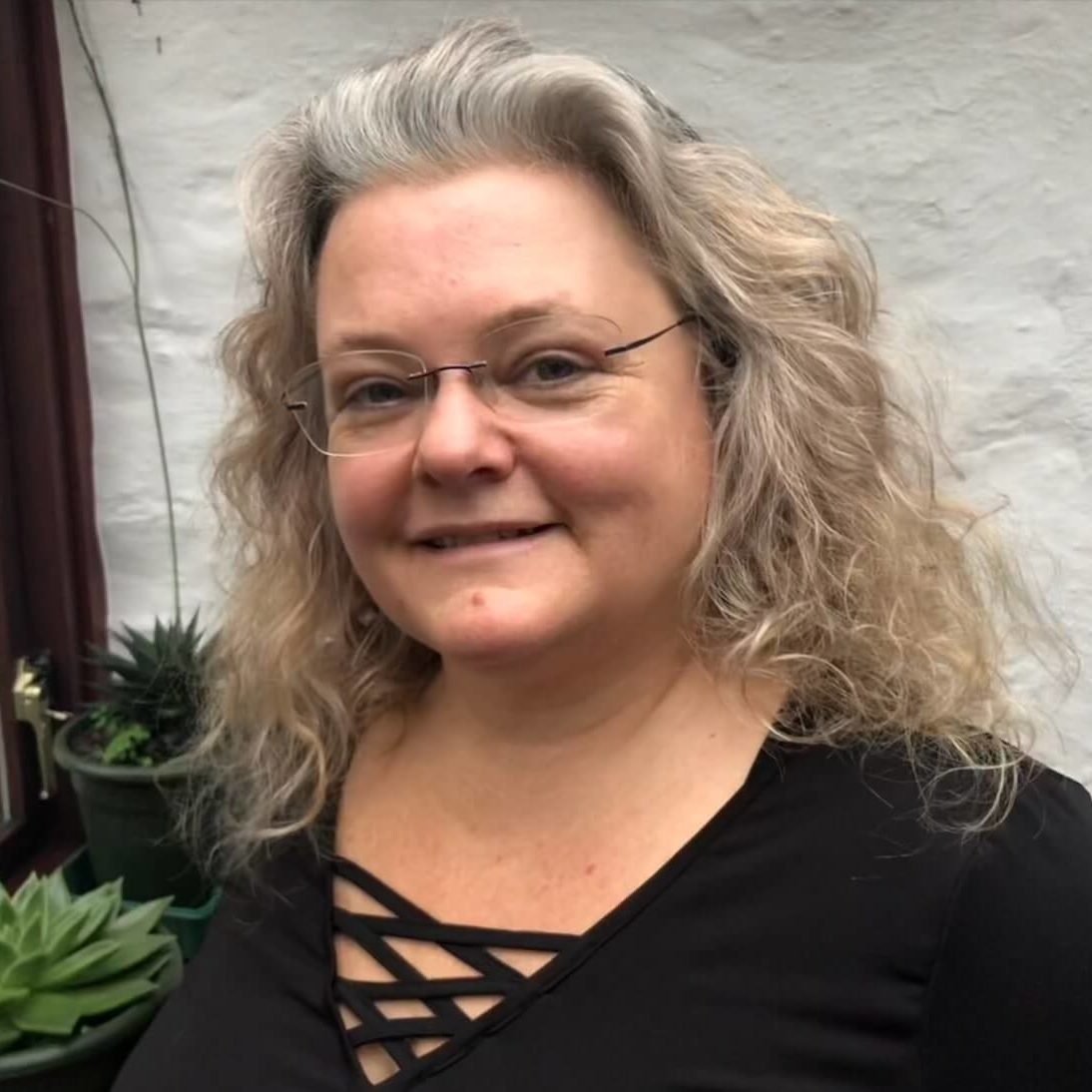 A woman with curly gray hair, glasses, and a black top standing next to green potted plants in front of a white brick wall.