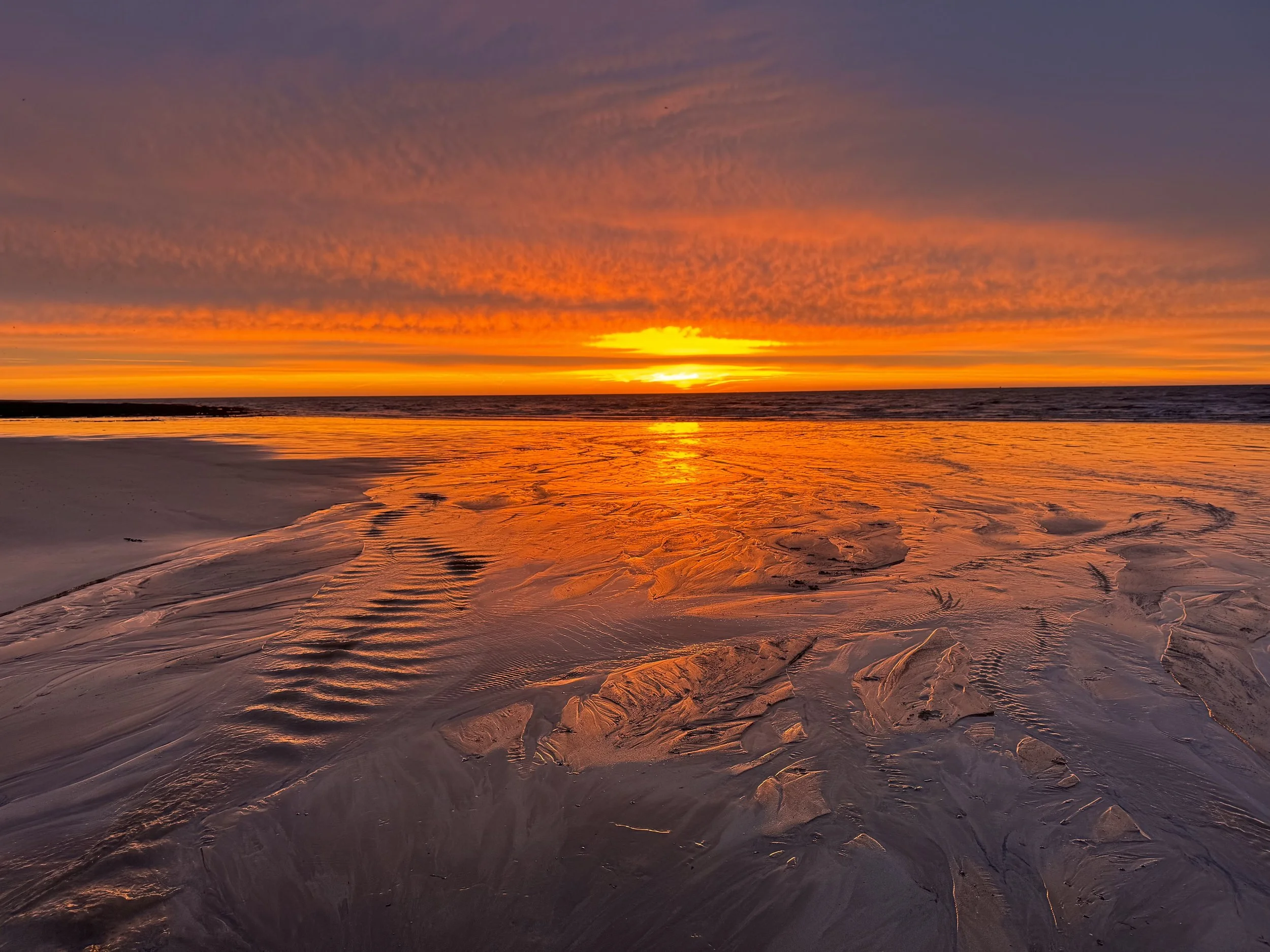 A vibrant sunset over a sandy beach with rippled sand and ocean waves in the background.