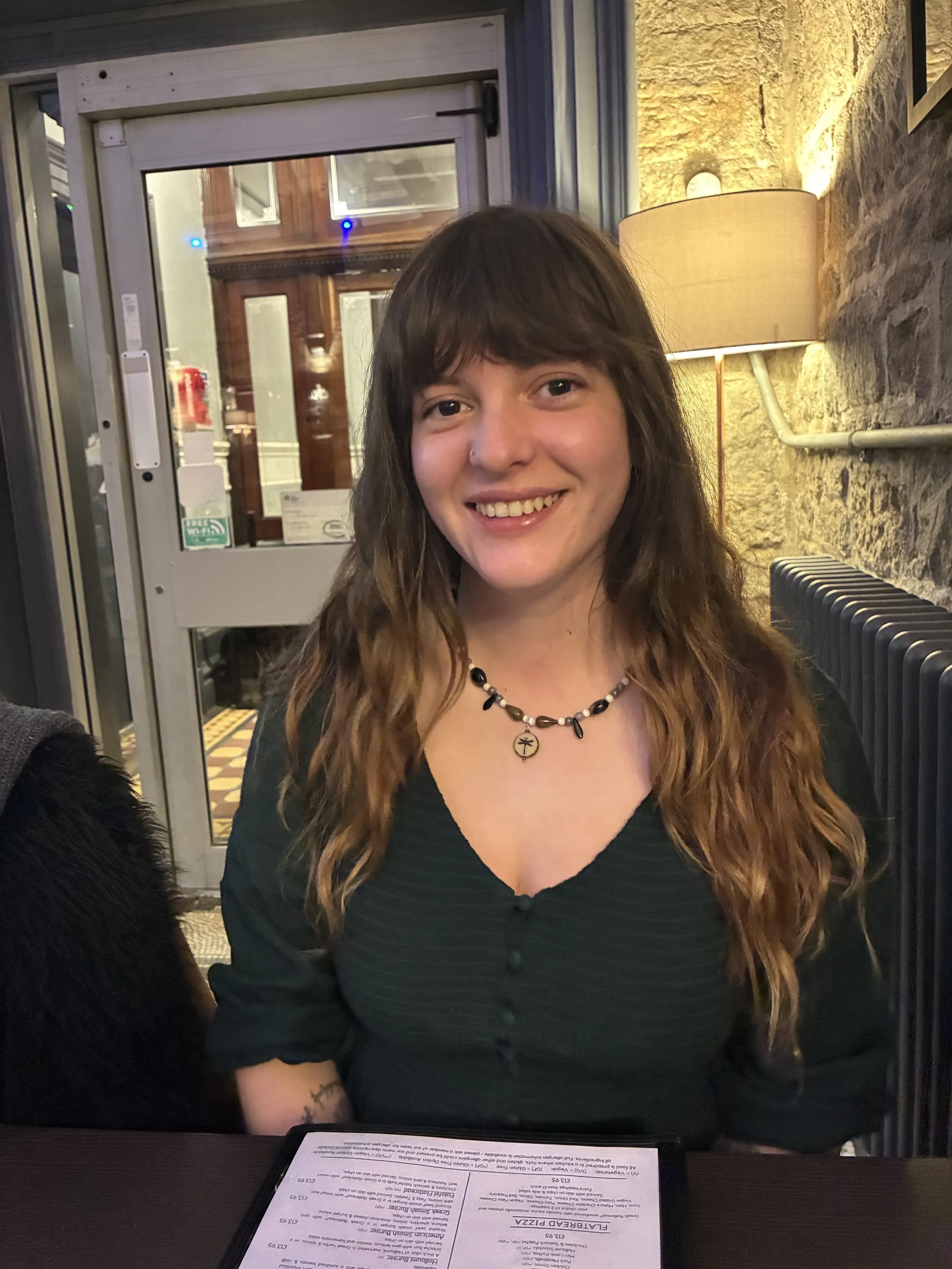A young woman with long wavy hair, wearing a black V-neck top and a beaded necklace, smiling while sitting at a restaurant table.