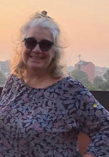 A woman with gray hair wearing sunglasses and a floral patterned top, standing outdoors with a city skyline and sunset in the background.