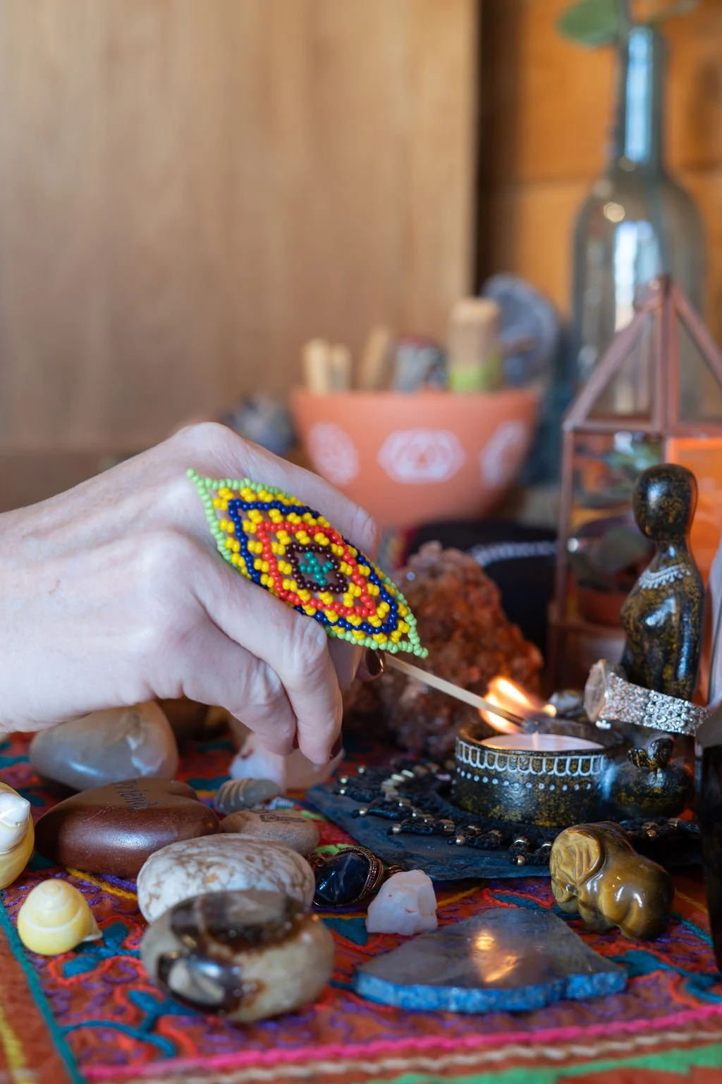 A person holding a beaded embroidery needlework in front of a table with various stones, jewelry, and decorative items, with candles and objects in the background.