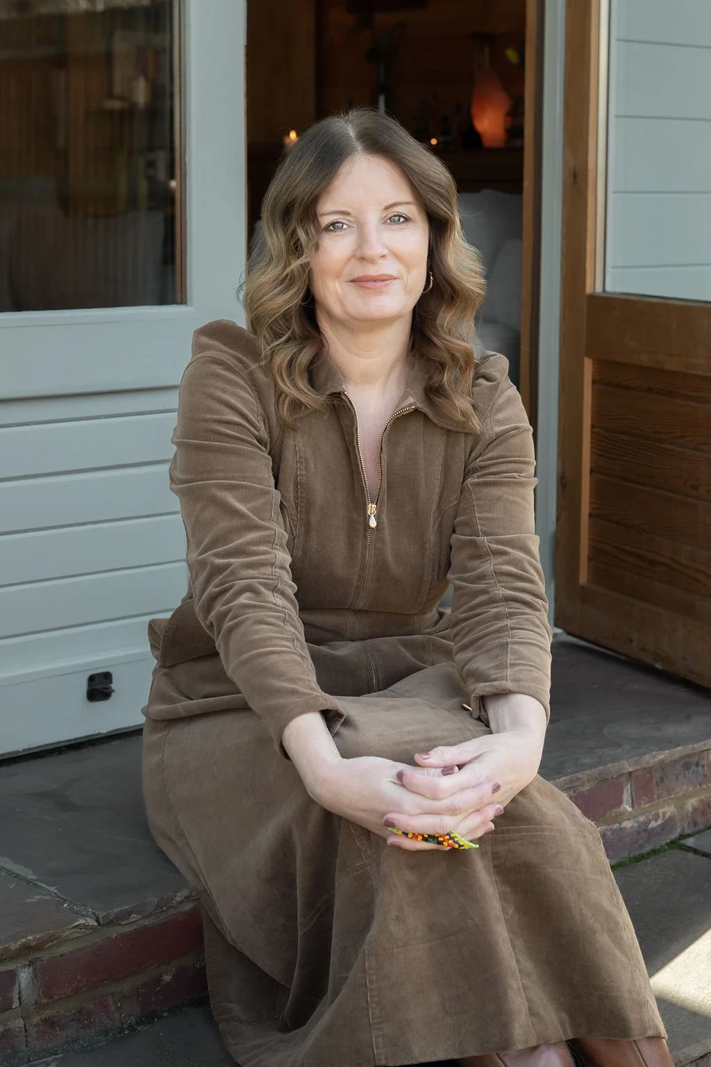 A woman with wavy, shoulder-length hair, wearing a brown zip-up corduroy jacket and matching skirt, sitting on a step outside a house.