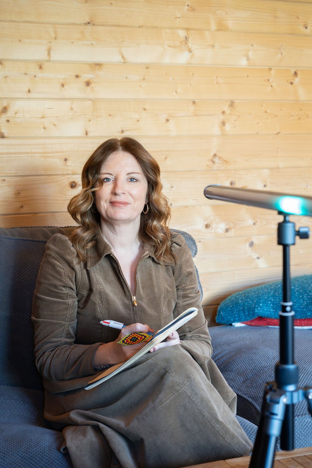 Woman sitting on a sofa, taking notes while being recorded by a camera with a wooden wall background.