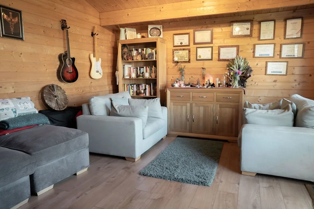 Cozy therapy room with wooden walls, featuring two sofas with cushions, a bookshelf filled with books, wall-mounted guitars, framed certificates or diplomas, and a sideboard with decorative items and a bouquet of flowers.