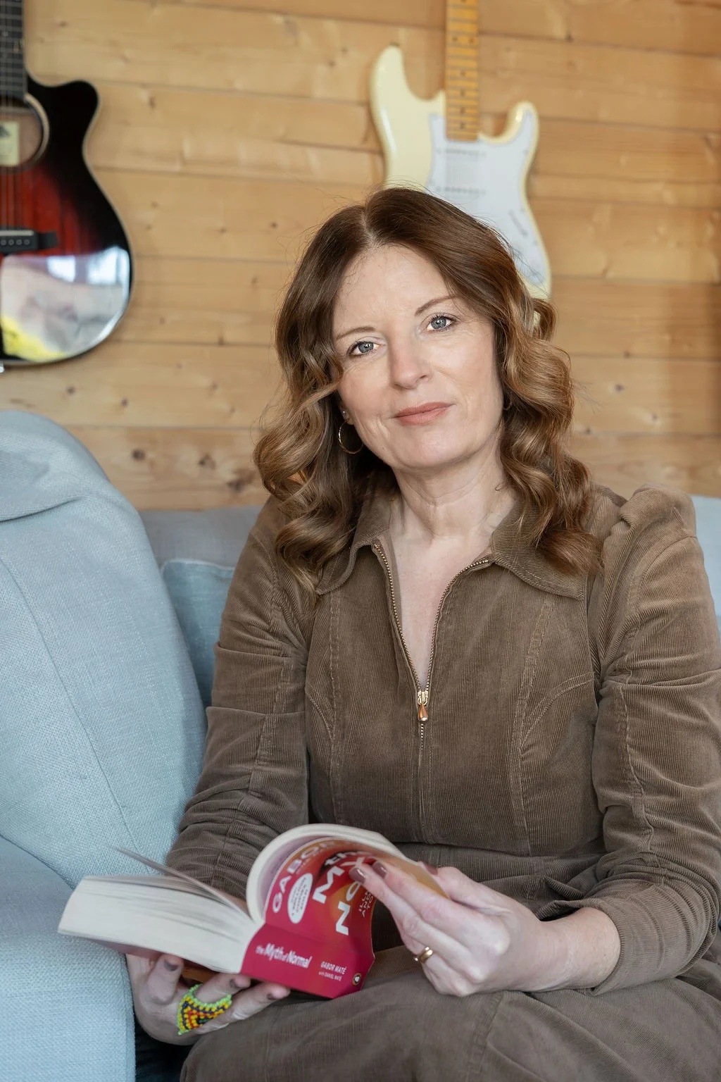A woman with wavy brown hair sits on a light blue couch in front of a wooden wall with musical guitars hanging on it. She is holding an open book or magazine and looking at the camera.