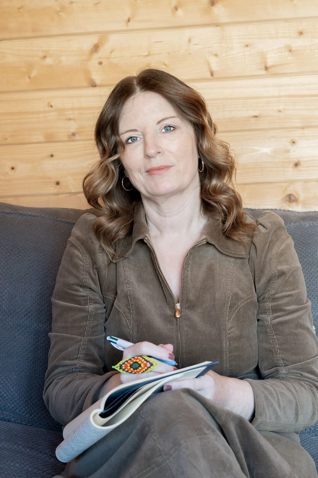A woman with wavy brown hair and blue eyes, wearing a brown zip-up jacket, sitting on a dark gray couch with a wooden wall background. She is holding a notebook and a pen, with some colorful beaded jewelry on her hand.