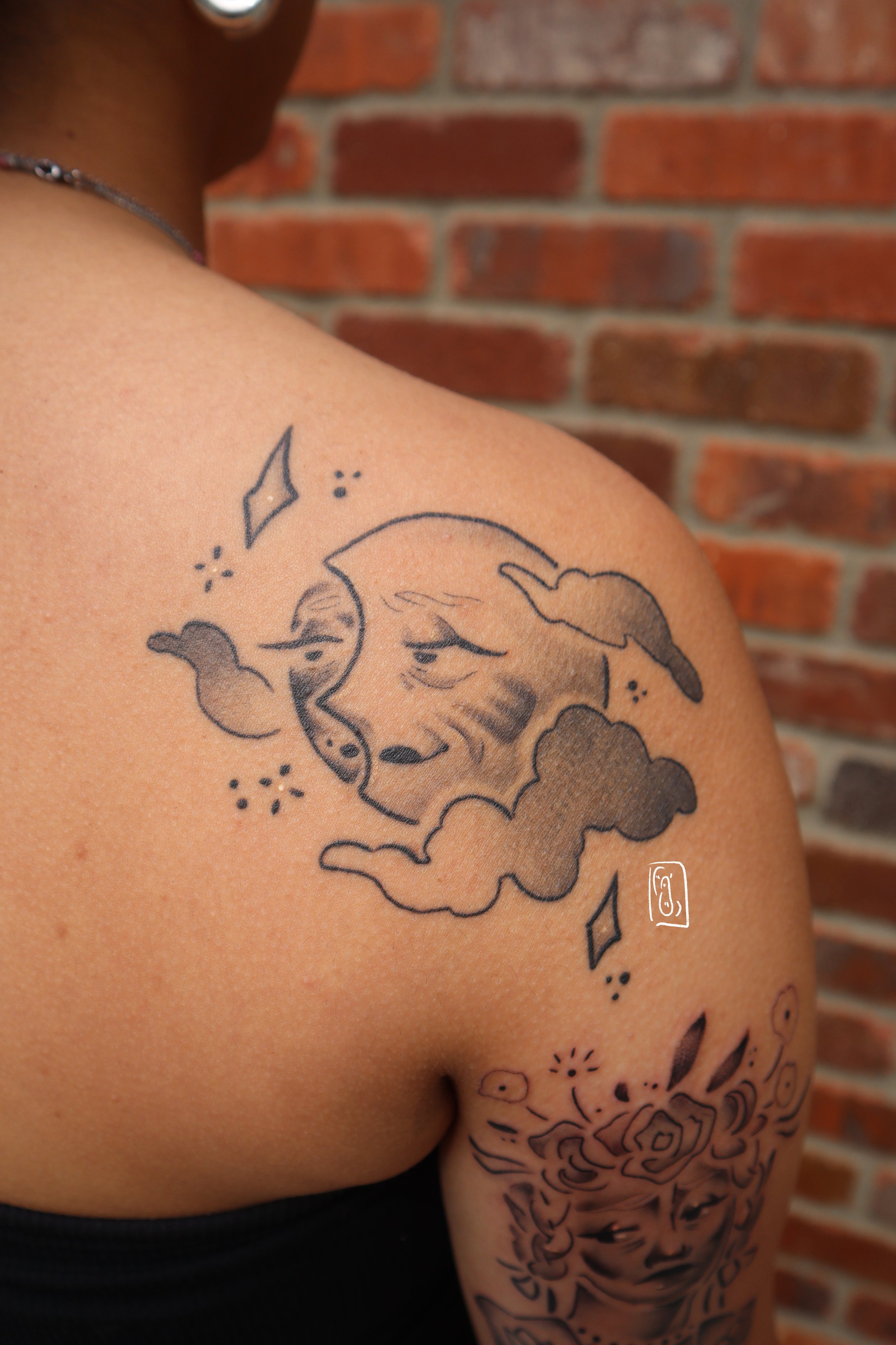 tattoo of a moon with a soft, aged, feminine face. it is on the shoulder blade of a person with more melanin in their skin and is black and grey. the tattoo is also healed and has no redness to it any longer