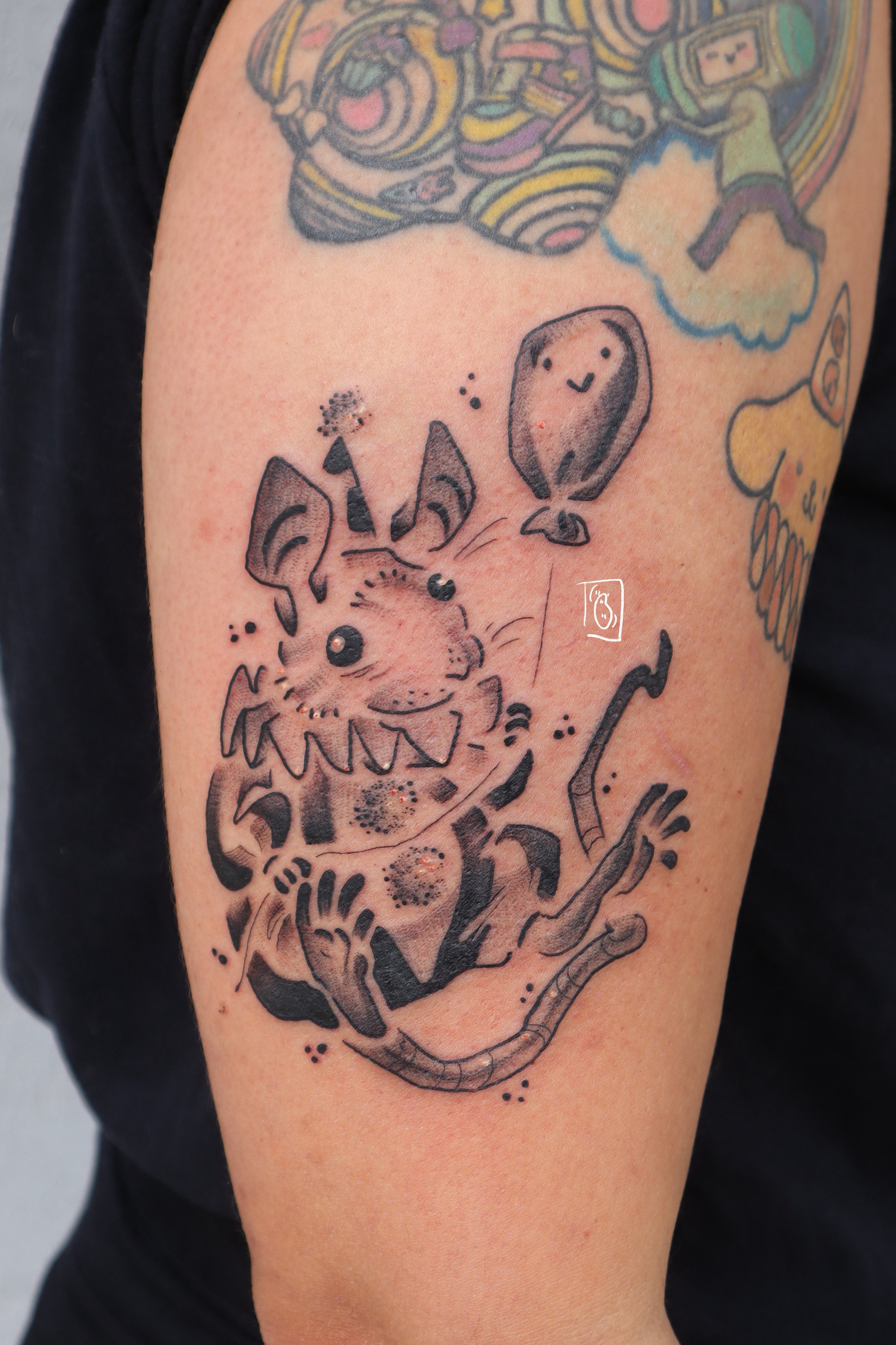 tattoo of a tiny rat in a clown costume. its eyes hold not a single thought behind them. he holds a balloon in his hands that has a smiling face on it. it is black and grey and on the client's outer arm above the elbow