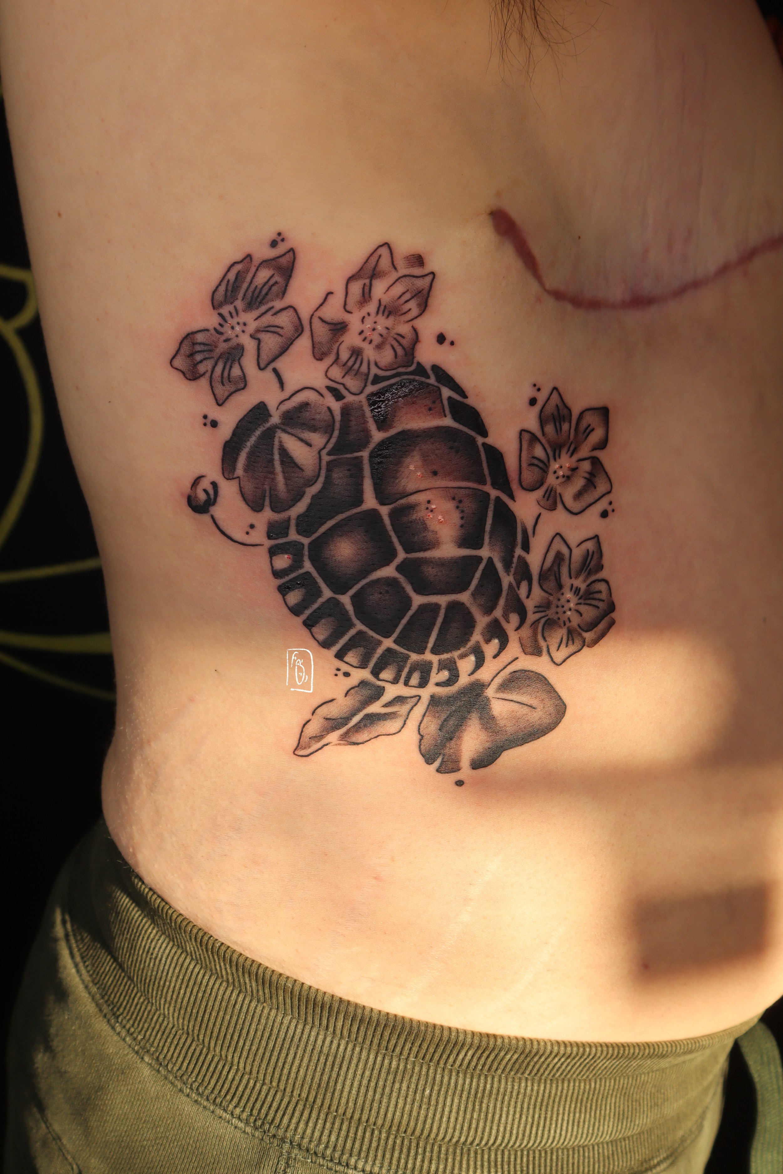 tattoo of a painted turtle's shell with marsh marigold flowers surrounding it. it is a black and grey piece on a person's side, and frames their top surgery scar beautifully