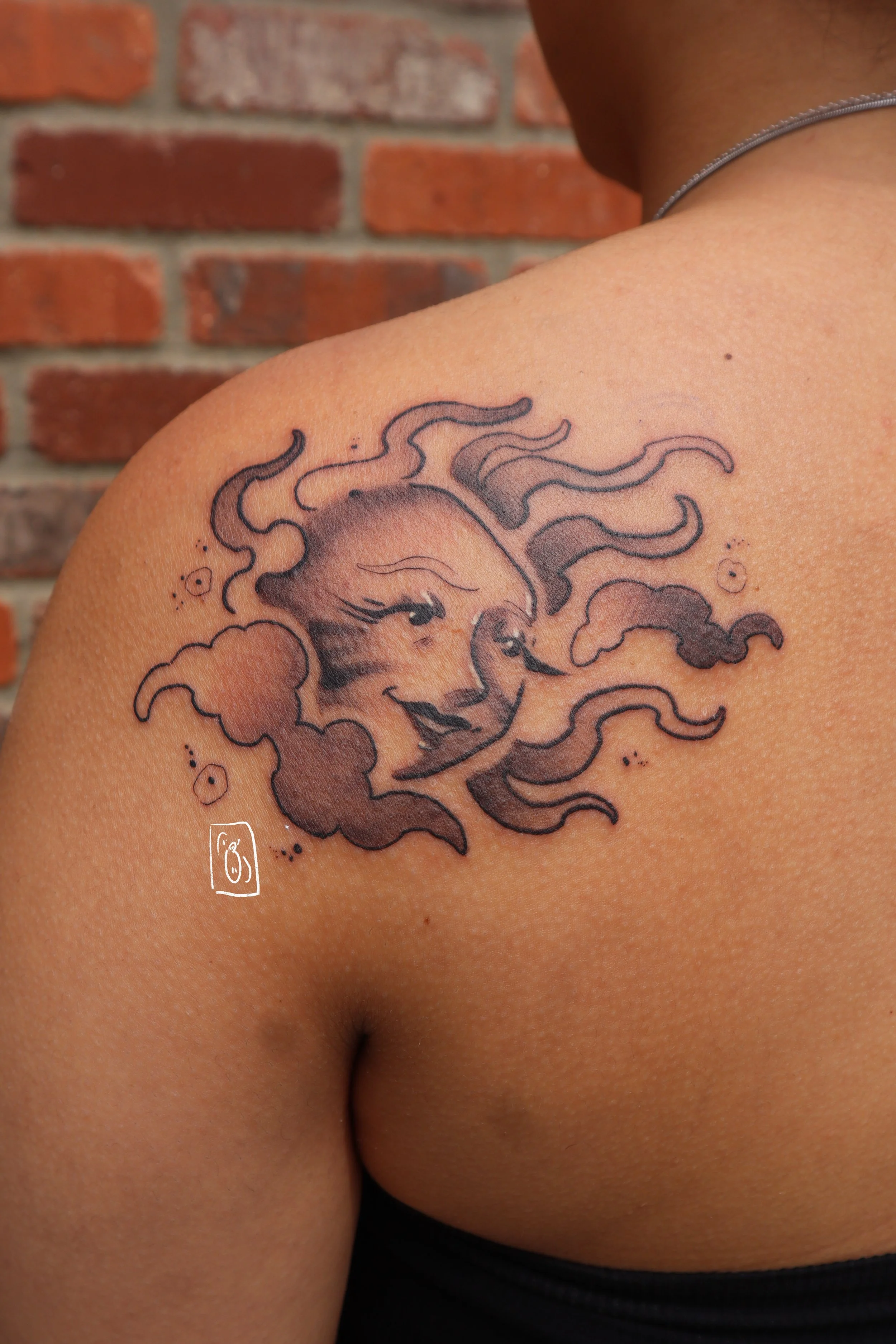 tattoo of a sun with a soft, feminine face. it is on the shoulder blade of a person with more melanin in their skin and is black and grey