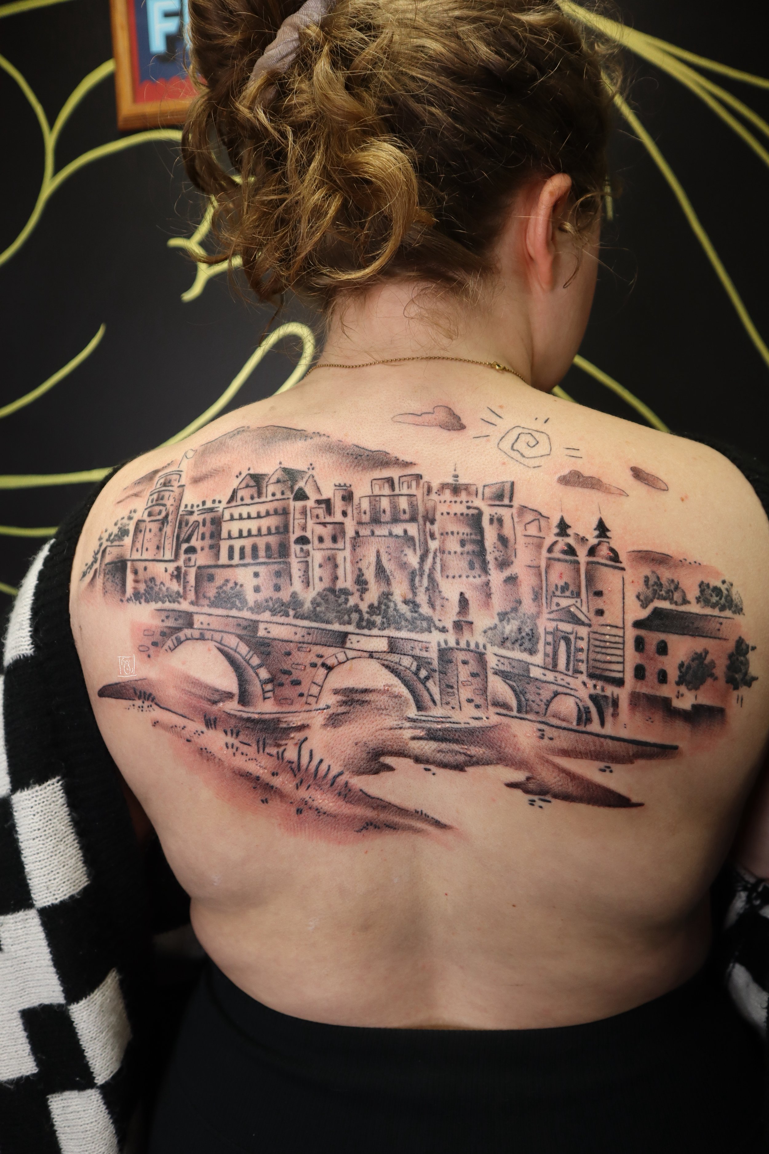 tattoo of an illustrative interpretation of the gate and city of hedeilberg, germany. the black and grey cityscape spans from shoulder to shoulder across the client's back