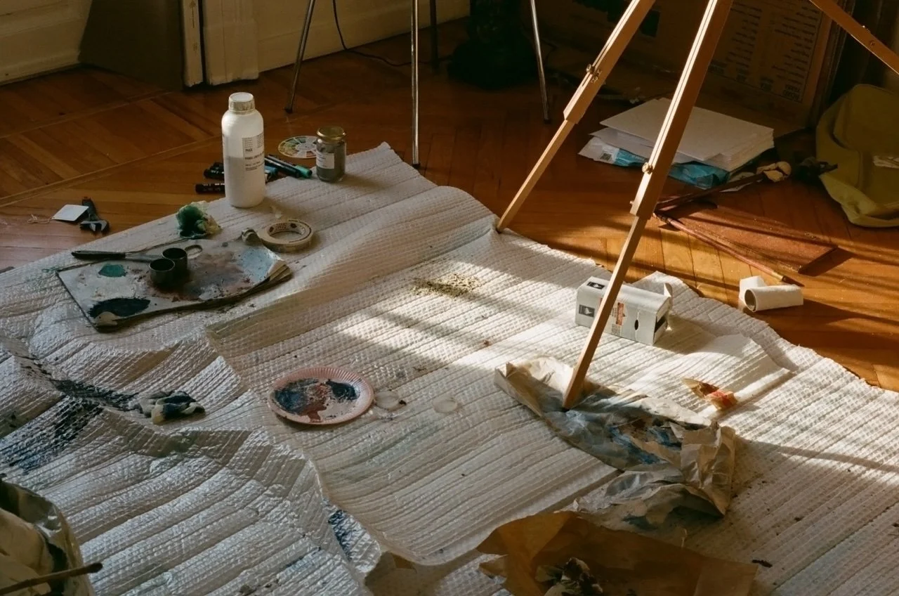 A room with mess indicating painting activities, including paint bottles, brushes, painter's tape, paint palettes with mixed colors, paper towels, and a paper plate with paint, with sunlight casting shadows.