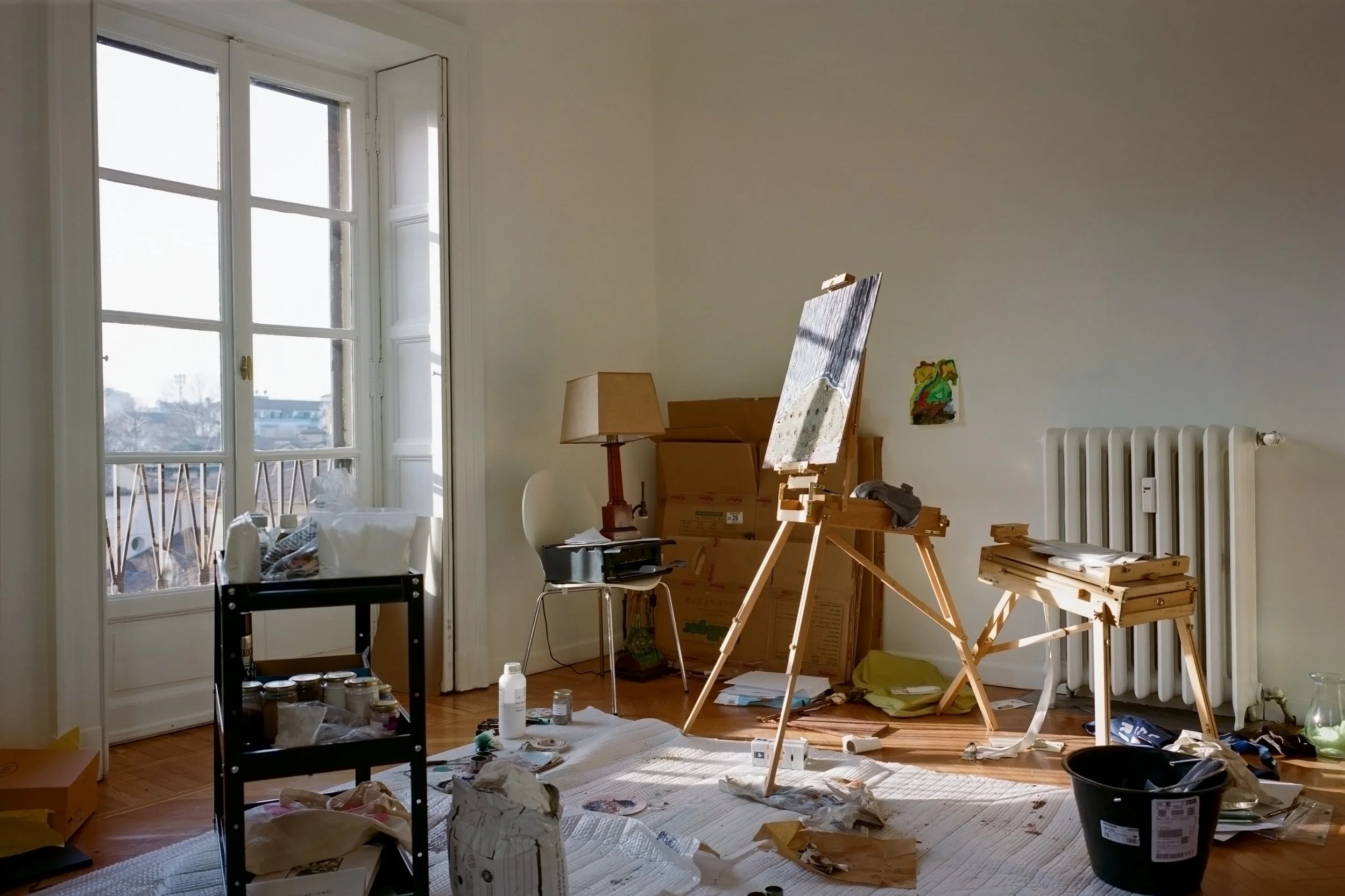 A cluttered room with art supplies and painting easels near a large window, with sunlight coming in, and various items scattered across the floor and surfaces.