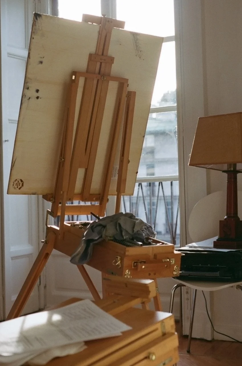 An artist's easel with a blank canvas in a room with a large window, a desk with papers, a lamp, and some electronic equipment.