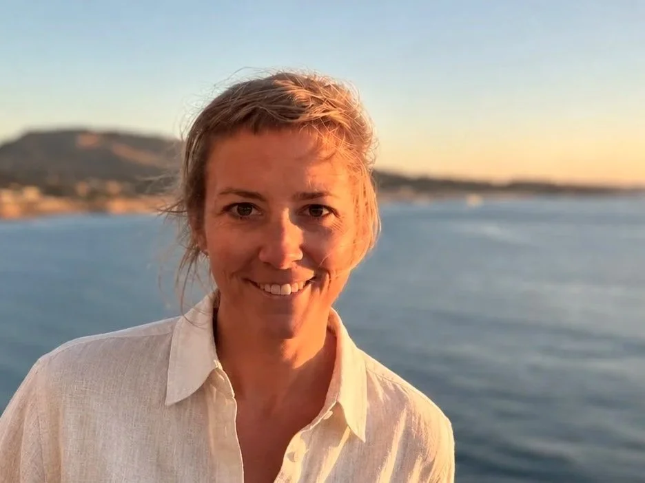 Dr. med. Dipl. mus Vanessa Eylers mit einladendem lächeln als Zeichen des Willlkommensheissen der PatientInnen , auch MusikerInnen, vor Wasser bei Sonnenuntergang, im Hintergrund ein Hügel als Zeichen dessen was gemeinsam zu überwinden ist - Nur Mut!