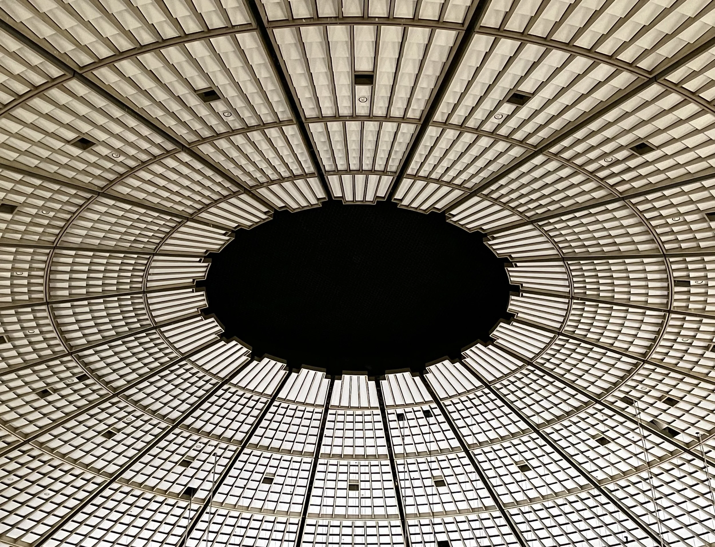 Innenaufnahme eines kreisförmigen Gebäudes mit einem hohen, gewölbten Dach aus Metall und Glas, das ein spiralförmiges Muster bildet.
