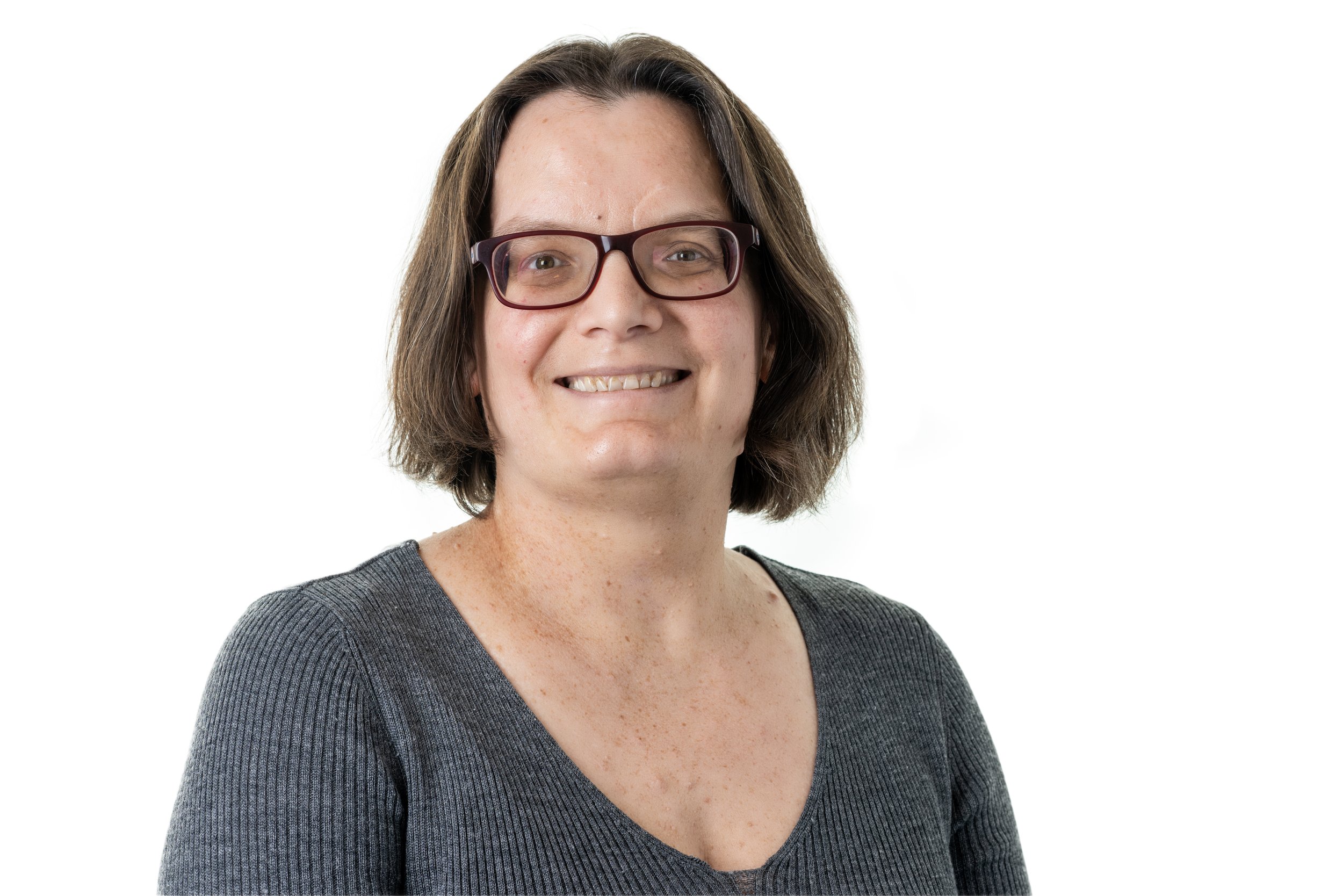 A woman smiling, wearing glasses and a gray top, with shoulder-length brown hair, against a white background.