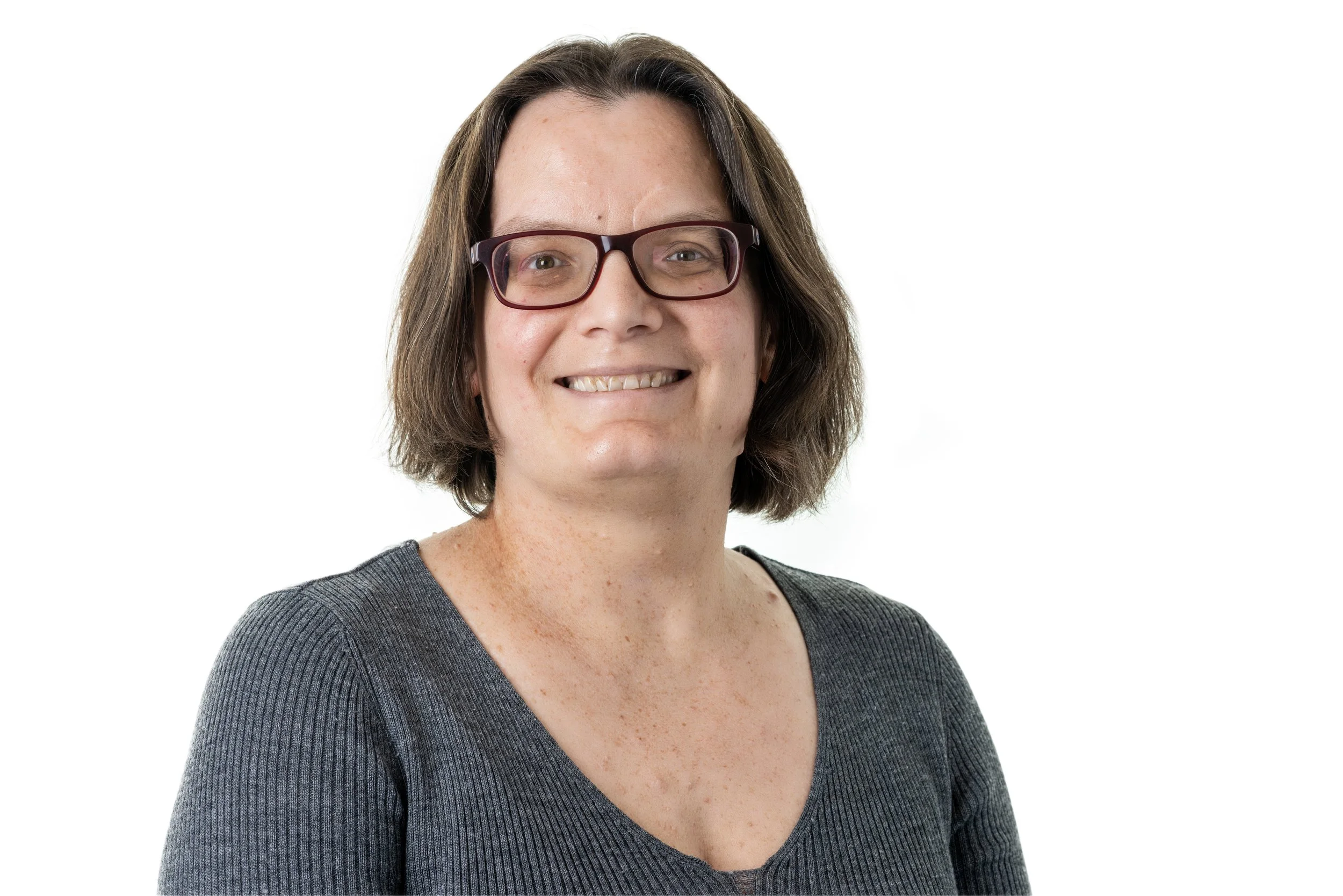 A woman with glasses and short brown hair smiling at the camera, wearing a gray top, with a plain white background.