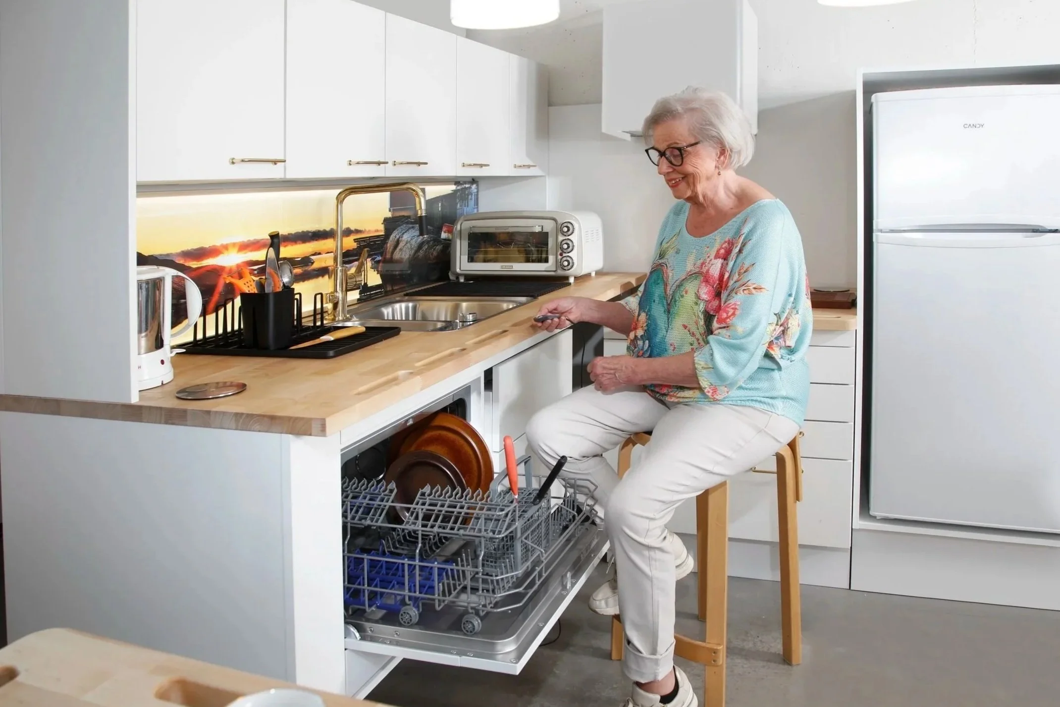 Iäkäs nainen istuu keittiössä tuolilla, tiskirattaat ja astiat avaavat astianpesukoneen.