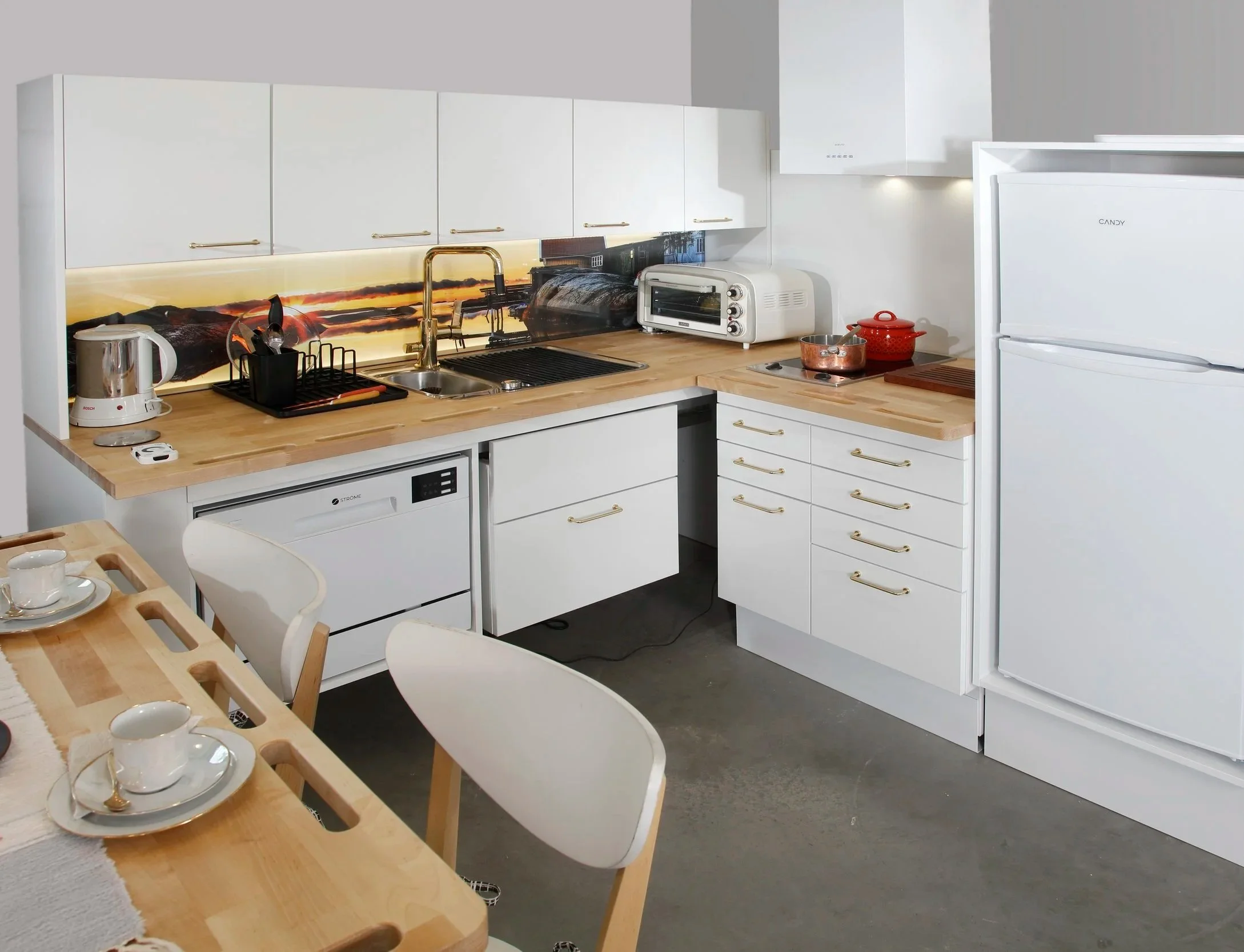 Valkoinen keittiö, jossa on puinen työskentelypöytä ja valkoisia kaappeja. Keittiössä on uuni, jääkaappi, mikroaaltouuni ja kahvinkeitin. Keittiön taustaseinässä on maisemamaalaus.