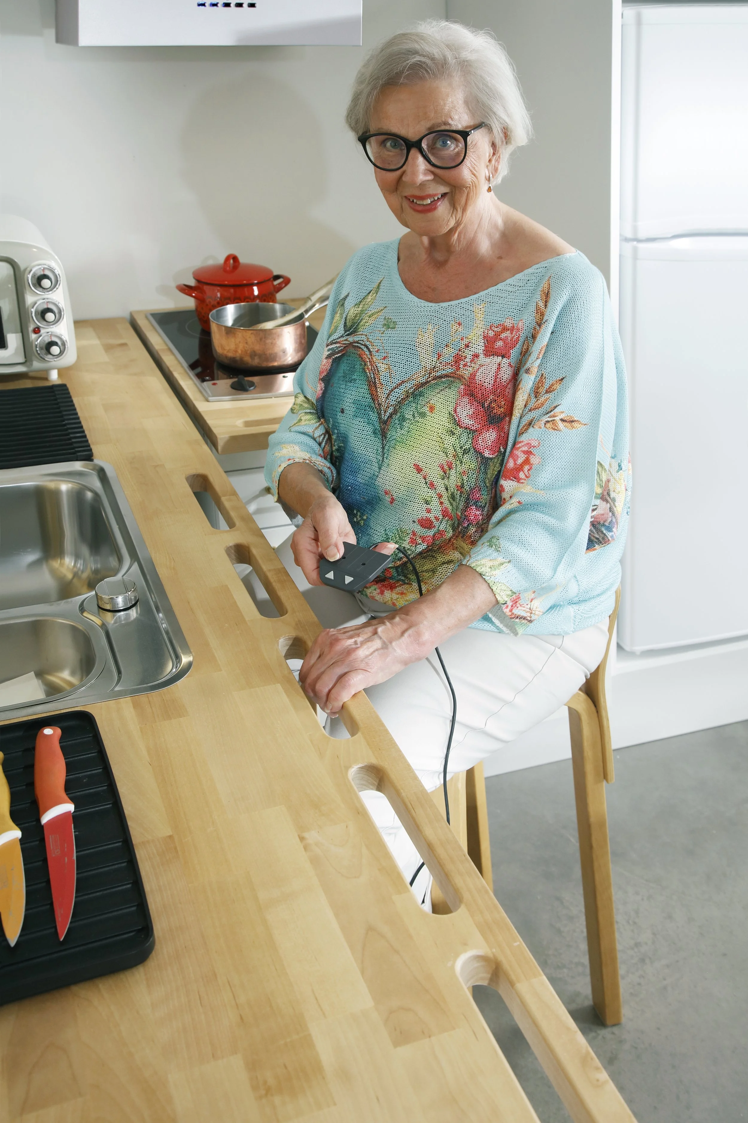Iäkäs nainen istuu keittiön baarijalalla ja käyttää kädessään kannettavaa hierontalaitepakettia, keittiössä on puhdas työpöytä ja keittiökoneita.