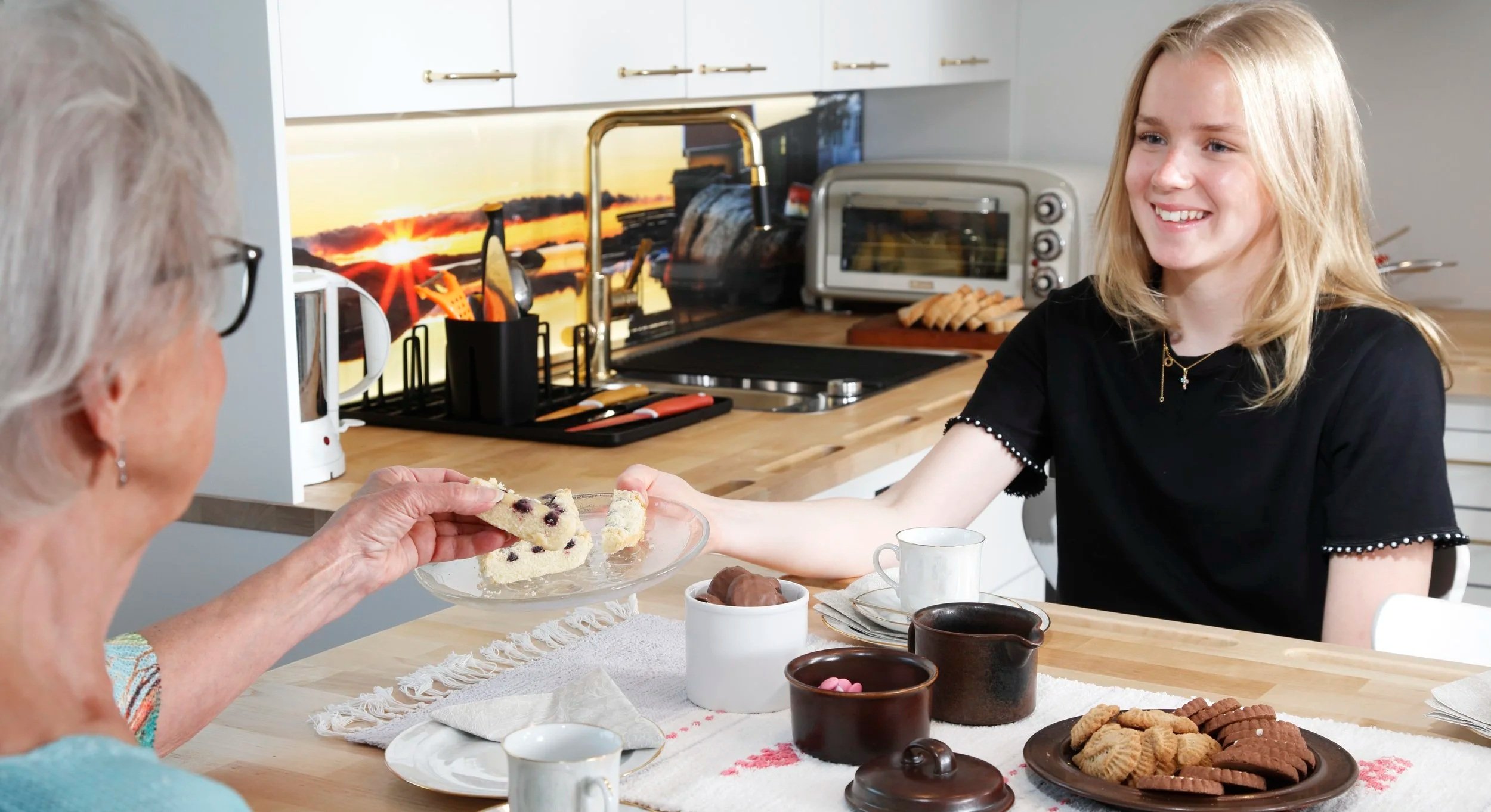 Nuori nainen antaa äidille tai isoäidille herkkupalan keittiössä, pöydässä on erilaisia herkkuja ja juomia.