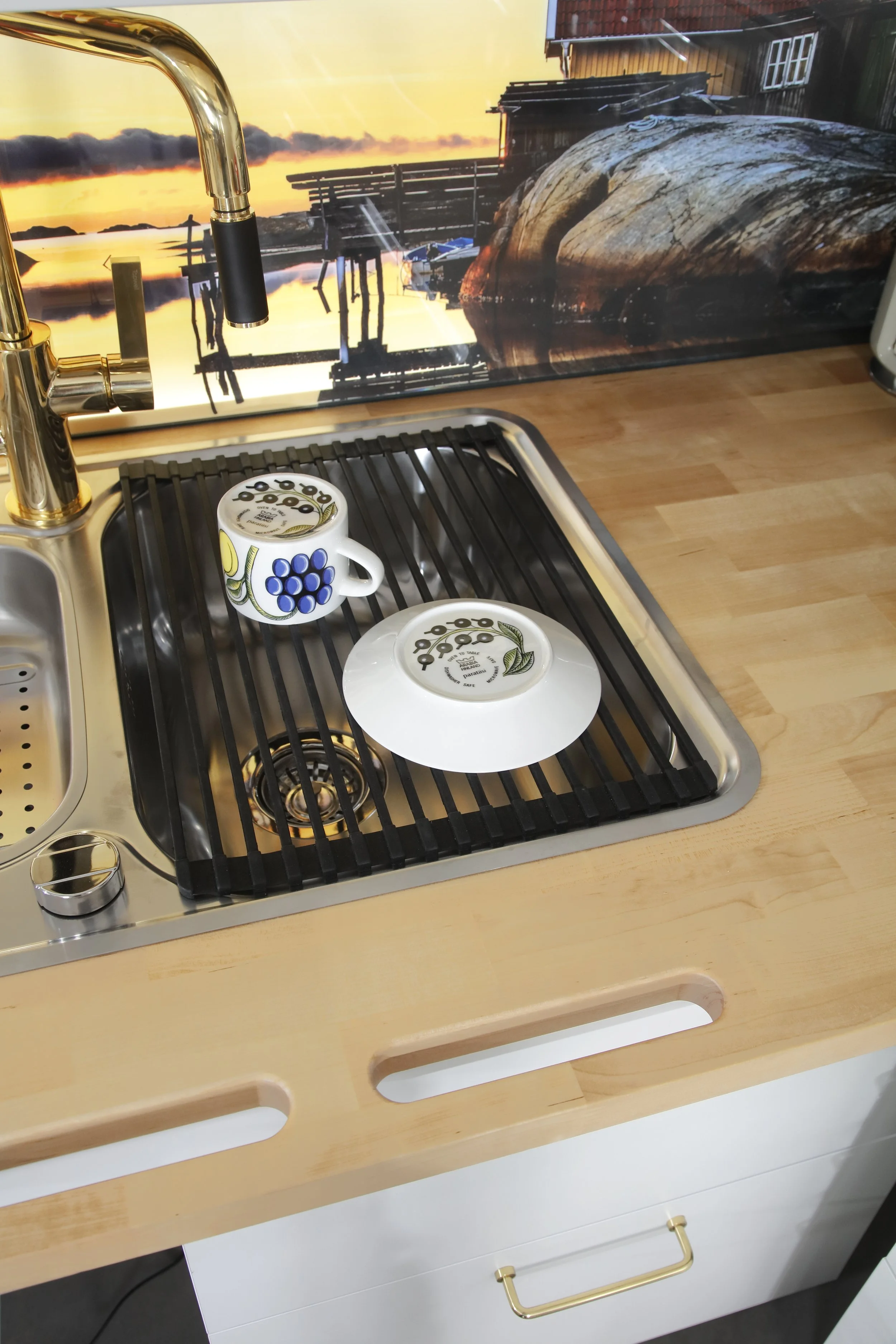 Keittöallas, jossa on kaksi kahvikuppia ja lautanen, sekä taustalla maisemakuva auringonlaskusta ja järvestä.