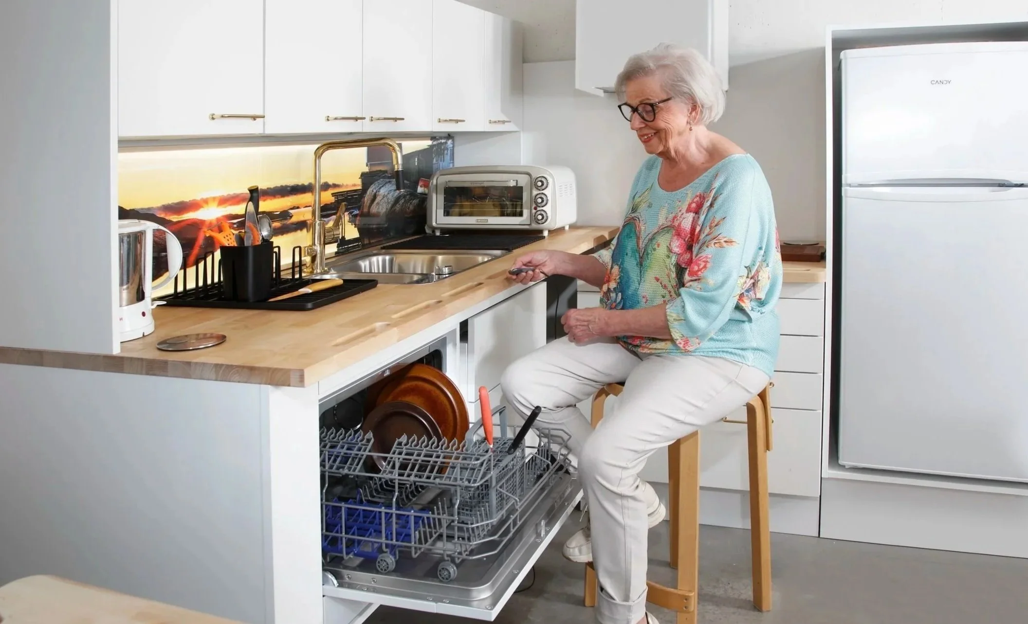 Nainen istuu jakkaralla ja ottaa tukea käsituista, jotta voi turvallisesti täyttää astianpesukoneen.