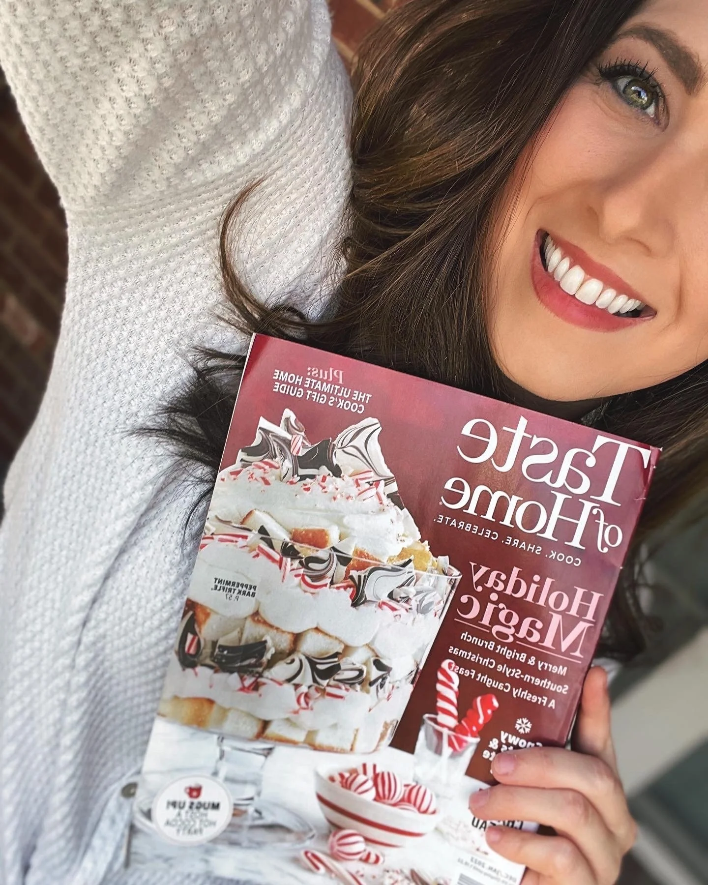 A woman smiling and holding a magazine featuring a holiday-themed dessert, with a close-up of her face and part of her arm visible.