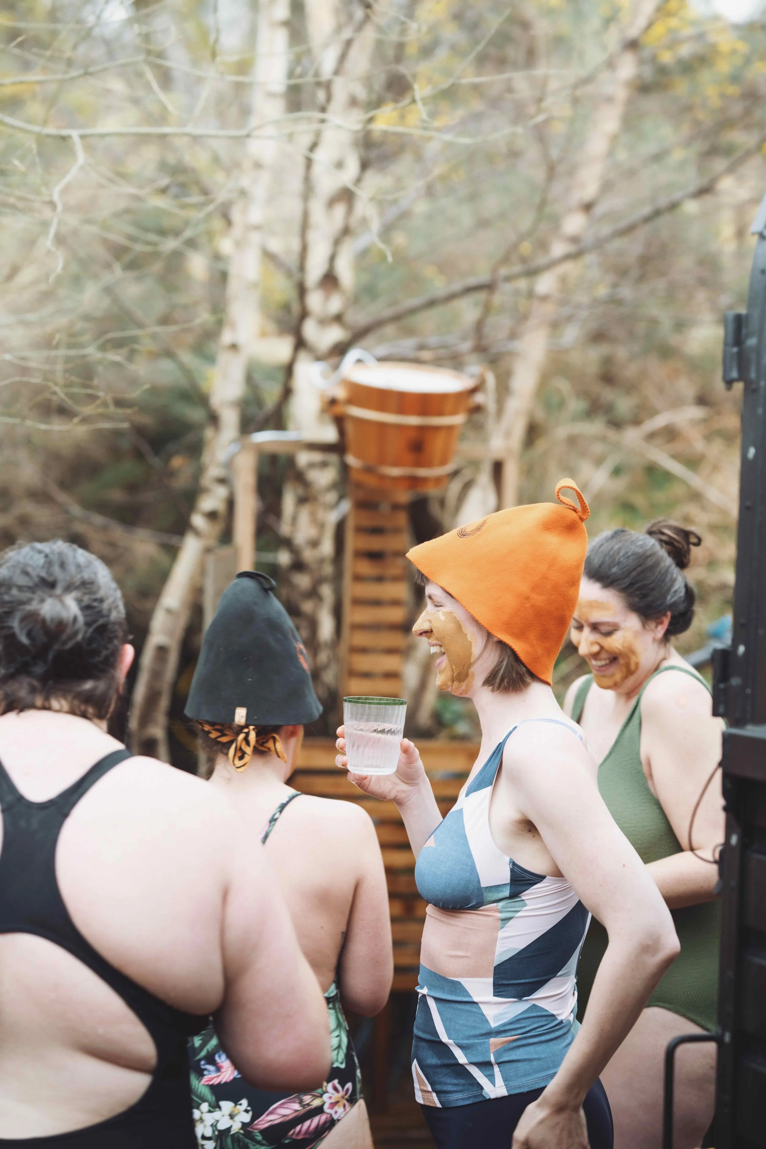 Sauna bathers stood outside. A woman is laughing, holding a drink and wearing a bright orange sauna hat