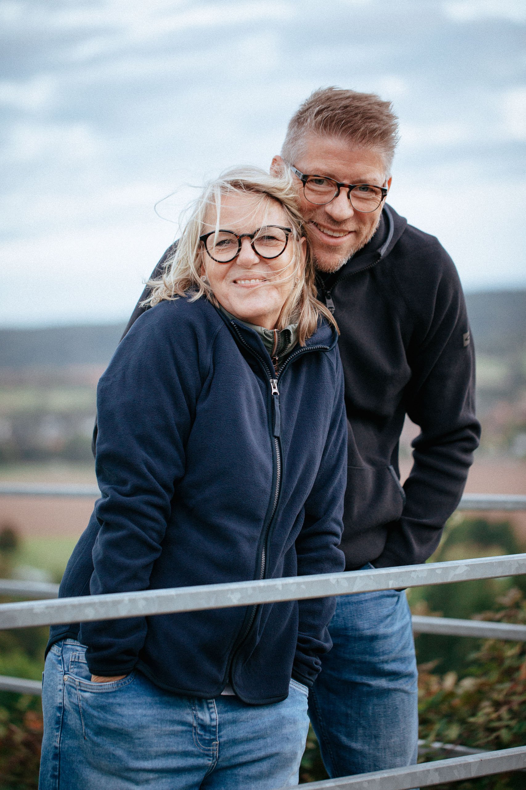 Ein lachendes Paar mit Brillen steht an einem Zaun im Freien, im Hintergrund sind weite Landschaften und Berge zu sehen.