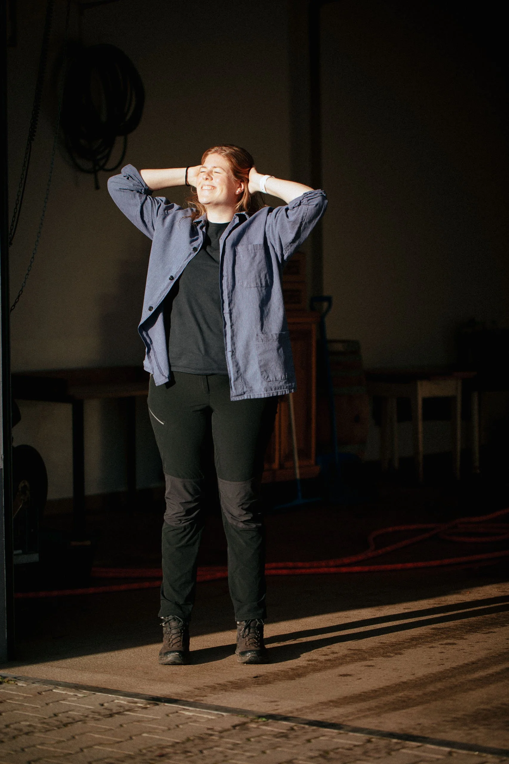 Eine lachende Frau steht in einem dunklen Raum mit Sonnenlicht, das auf den Boden und sie fällt, mit den Händen hinter dem Kopf, trägt eine blaue Jacke, schwarze Hose und Wanderschuhe.